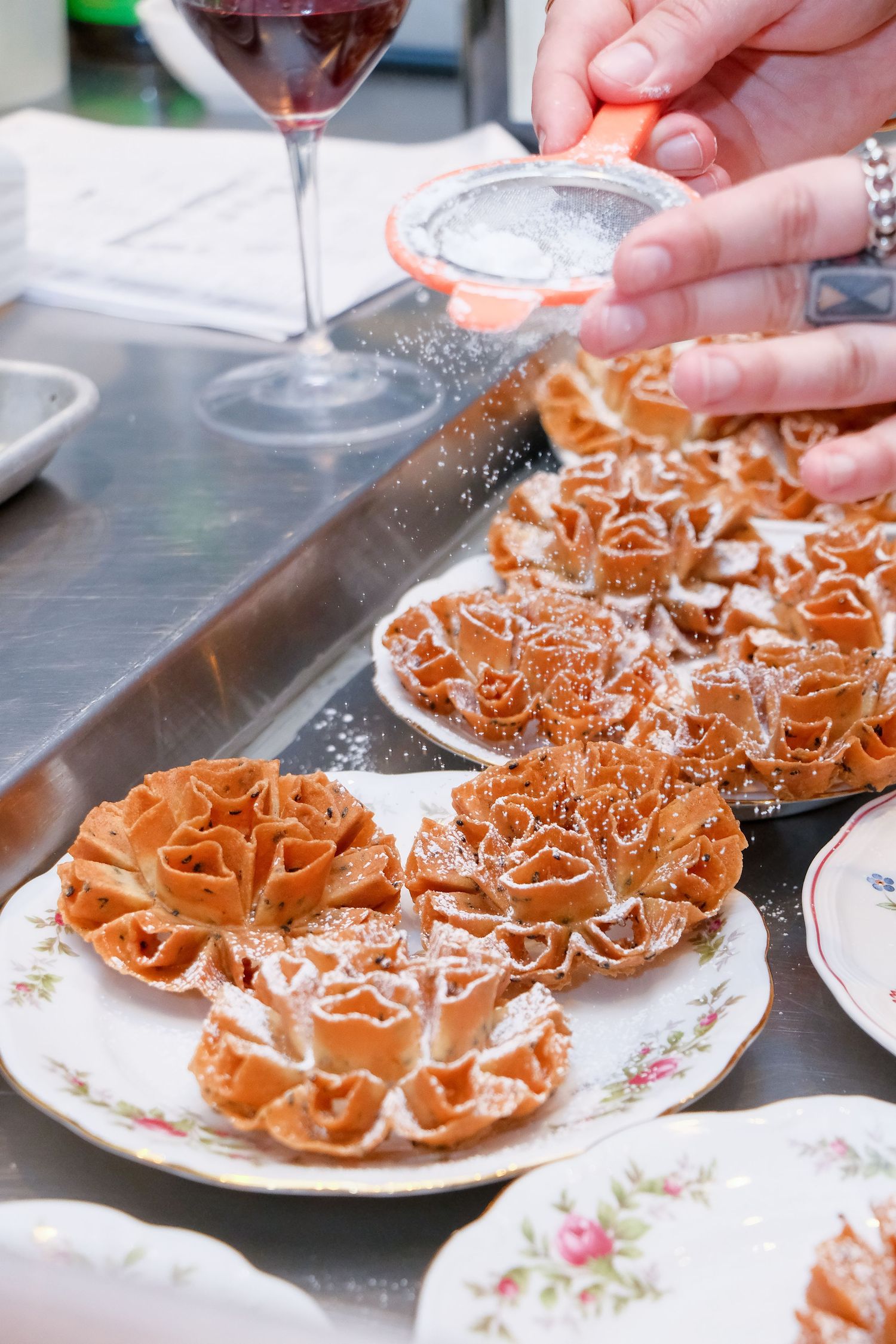 Hands dusting powdered sugar over golden rosette cookies on floral plates beside a glass of red wine.