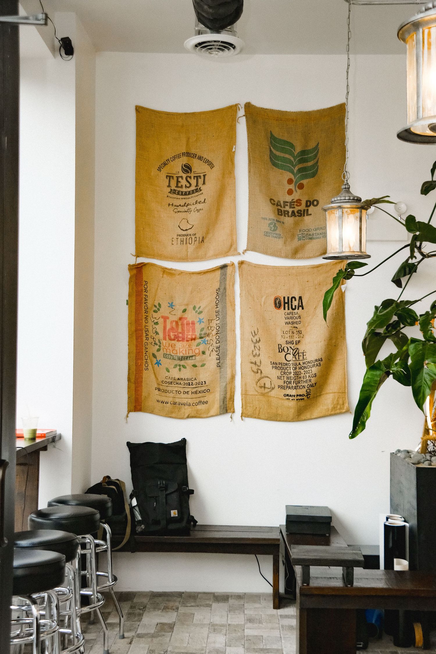 Rustic coffee shop interior with burlap coffee sacks on white wall, wooden bench, black backpack, and hanging lantern.