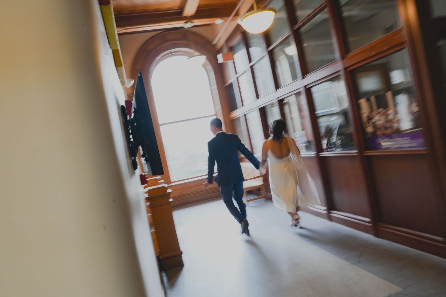 Bride and groom walking hand in hand down an elegant indoor hallway with arched windows on their wedding day.