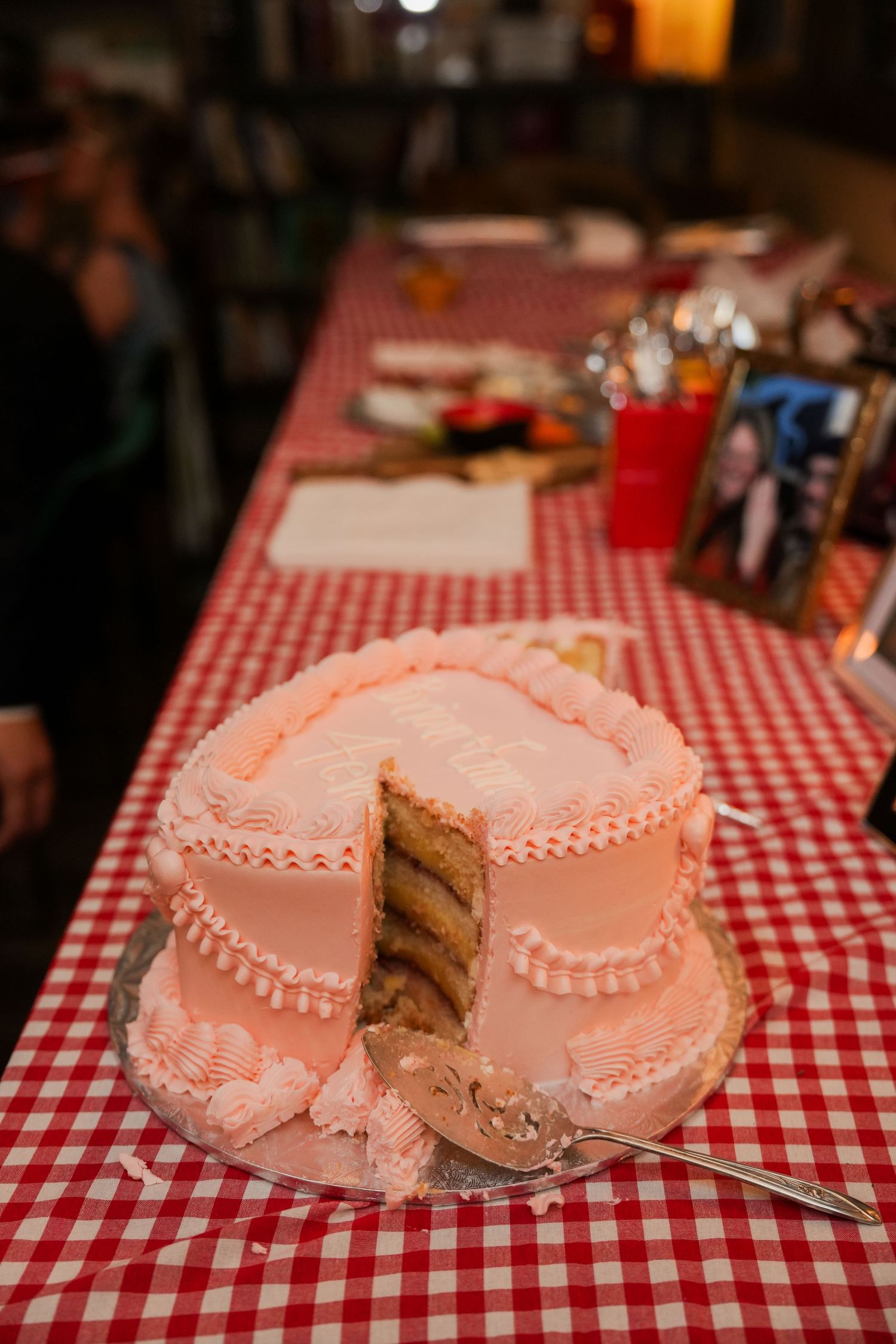 Pink frosted layer cake with a slice removed on a red checkered tablecloth at a party celebration.