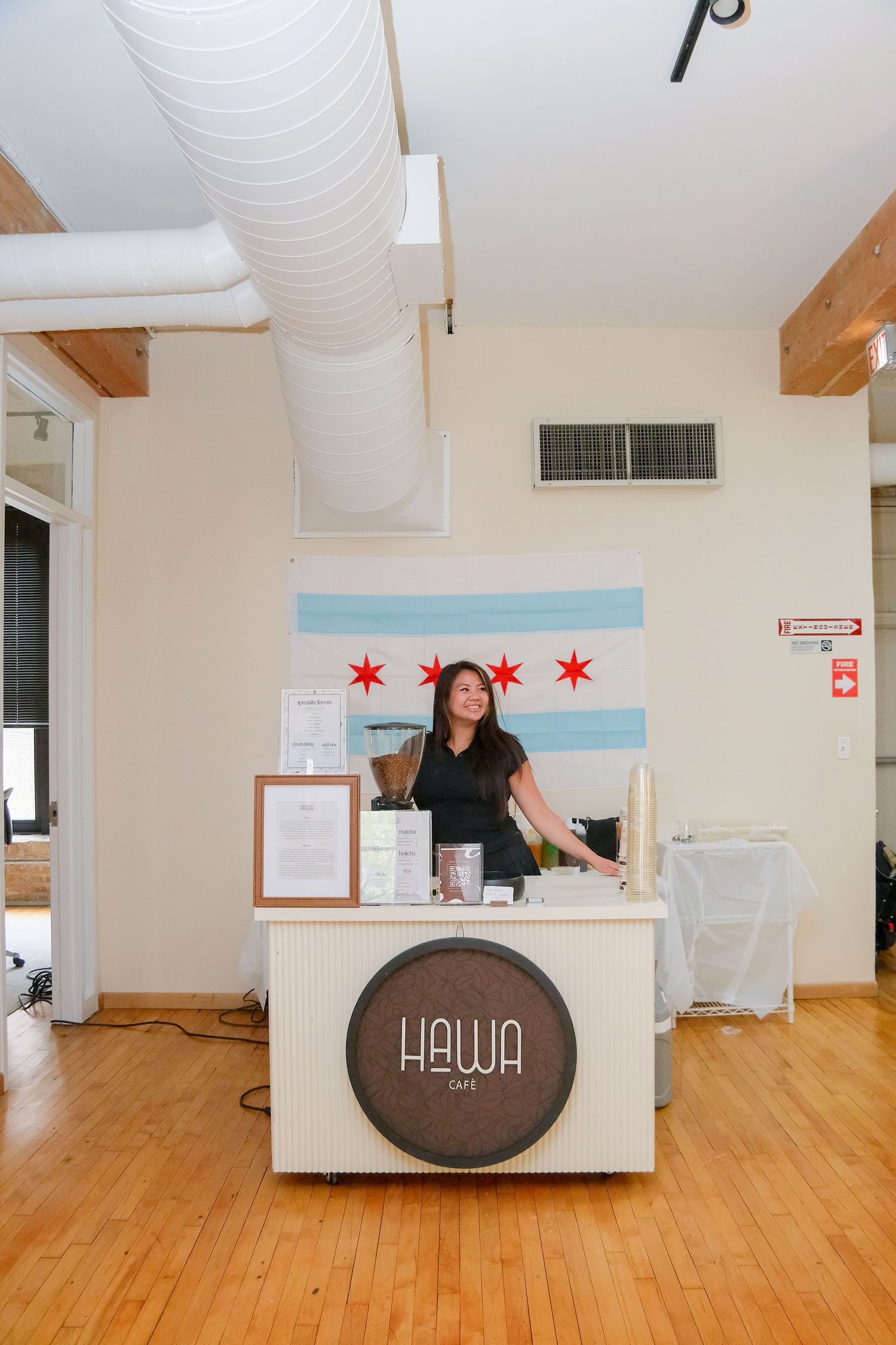 Woman standing behind a Hawa Cafe counter with Chicago flag backdrop in a bright, modern space with hardwood floors.