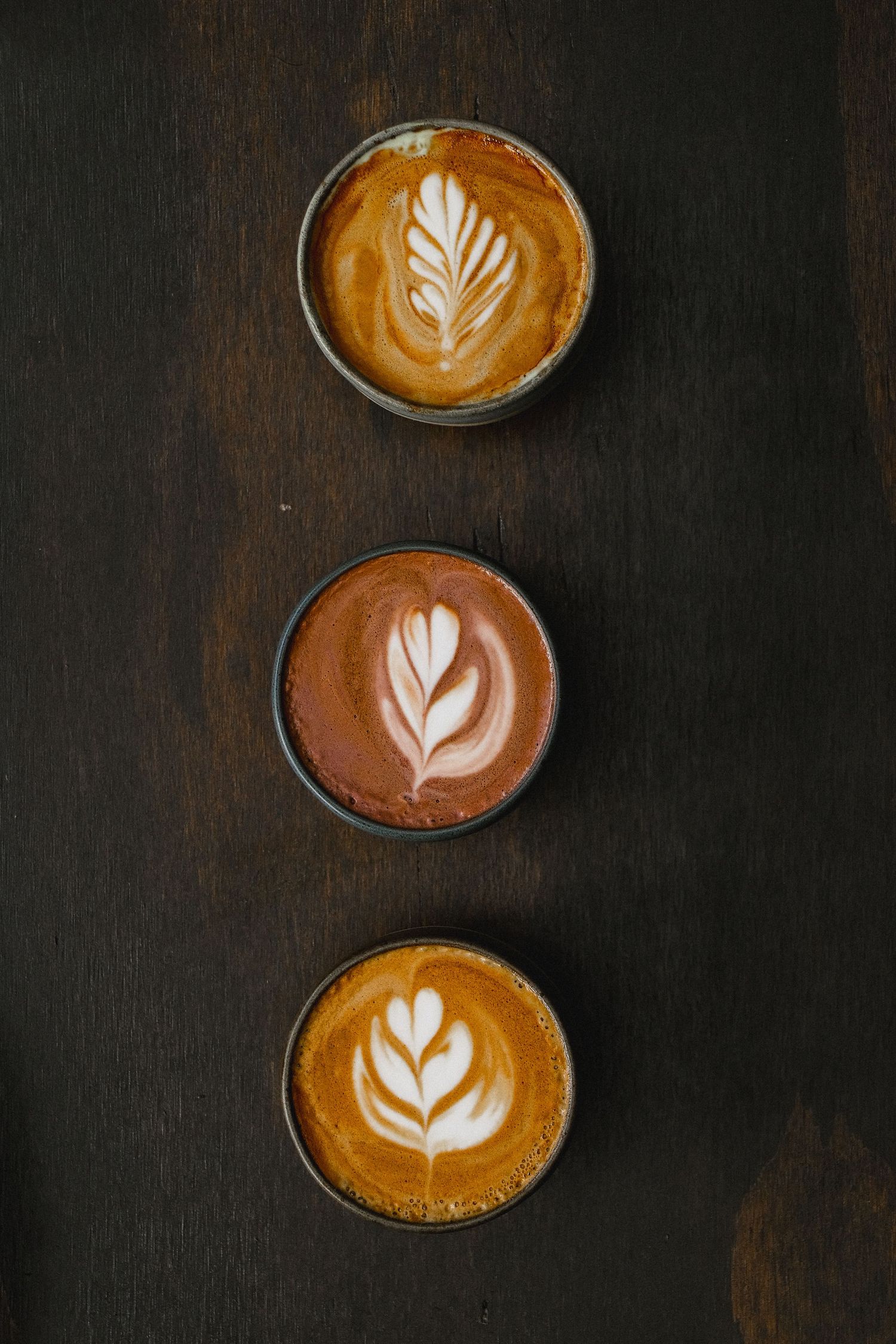 Three cups of artisanal coffee with intricate latte art designs arranged vertically on a dark wooden surface.