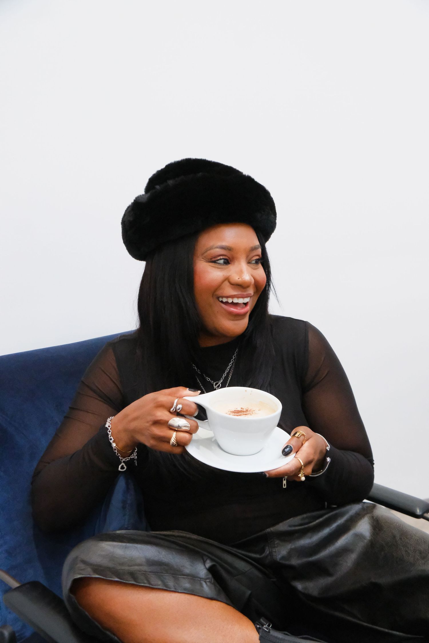 Smiling Black woman in black beret and outfit holding cappuccino cup while seated in blue chair.