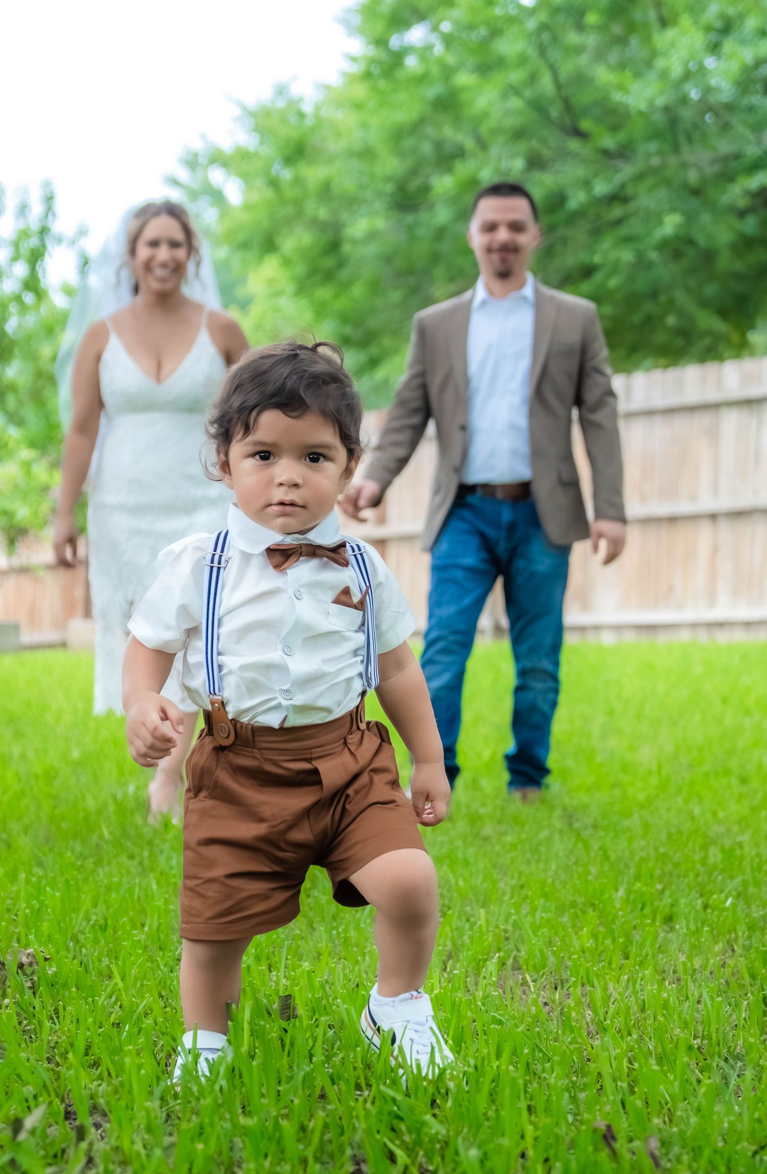 Small child in brown shorts and suspenders walks through green grass during wedding photos.