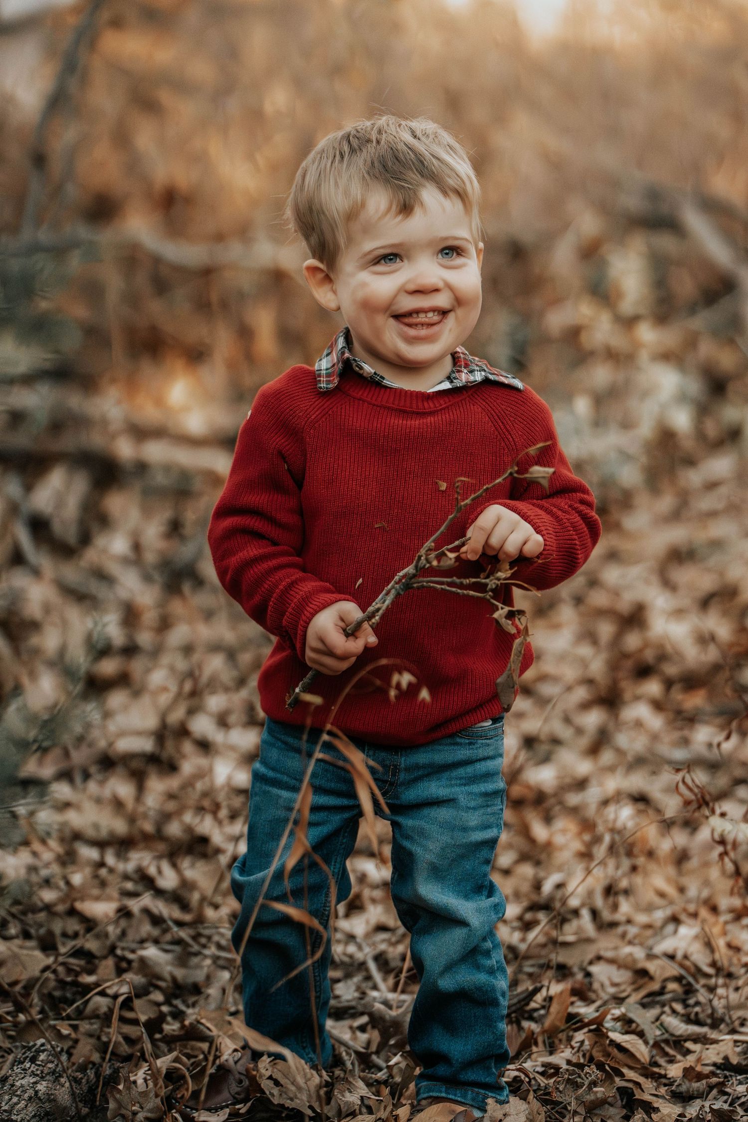 Young child in red sweater holding sticks and smiling in autumn forest setting.