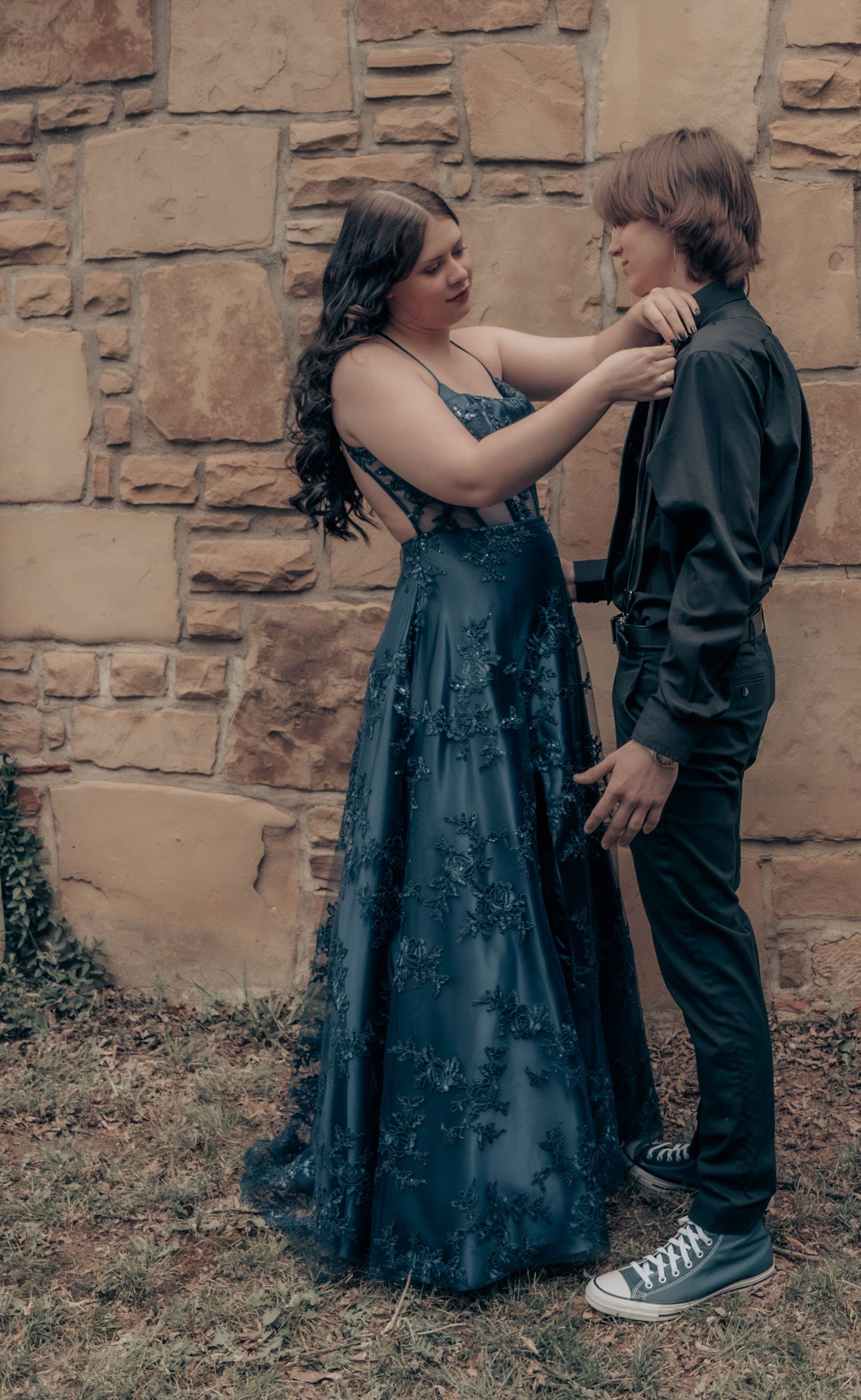 A formal couple poses against a stone wall backdrop with blue dress and black suit details.