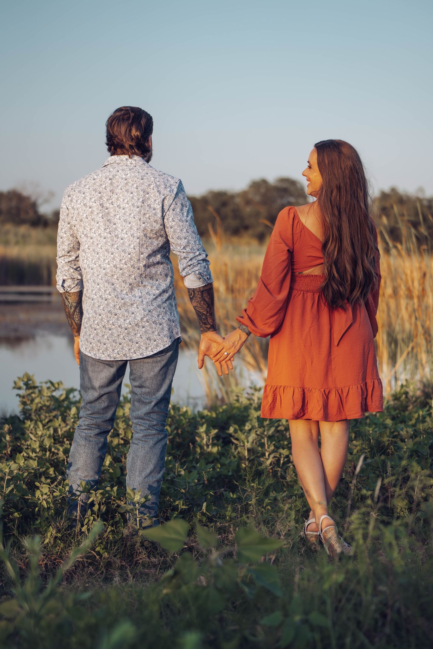 Two people walking through tall grasses and flowers beside a lake at sunset.
