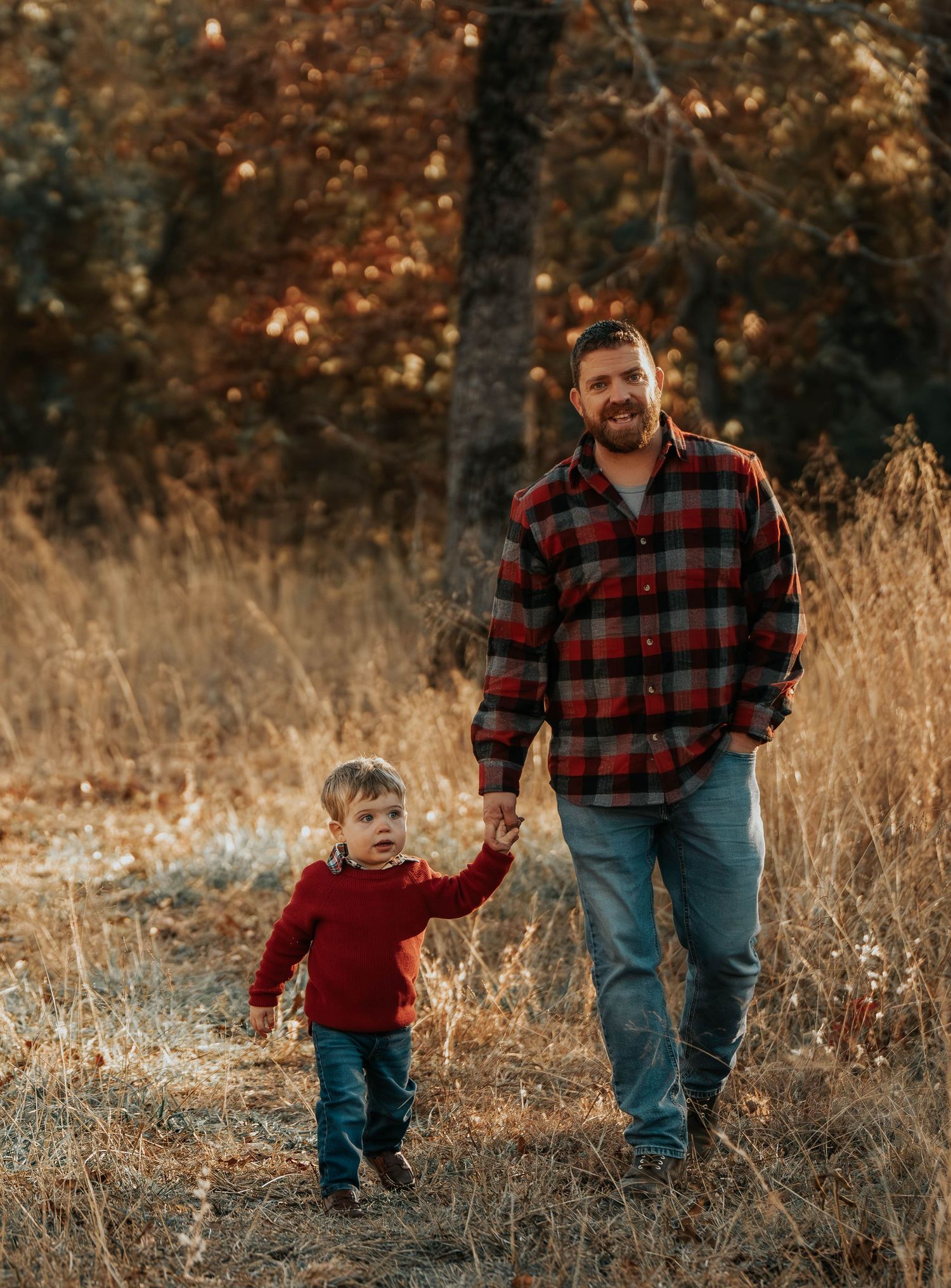 Adult and child walking hand in hand through tall grass in autumn forest.