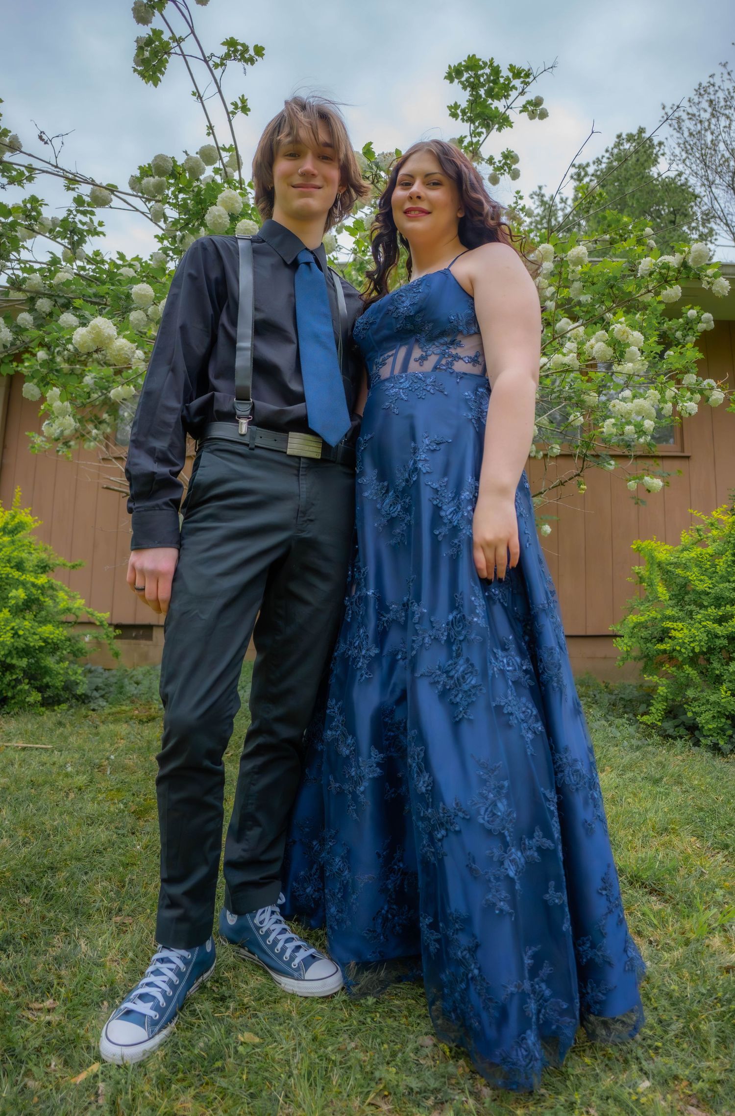 A couple poses in formal attire with a navy blue dress and dark suit against greenery and a wooden fence.