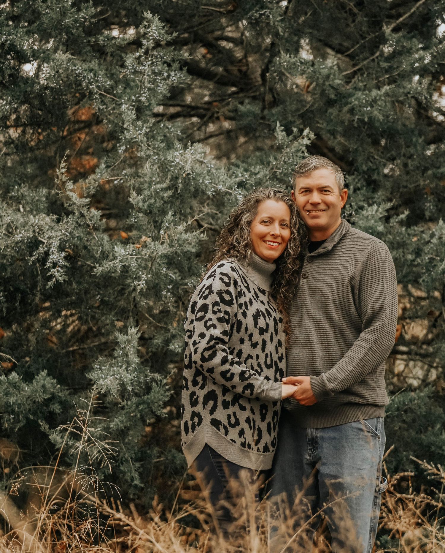 A couple in casual sweaters poses together against an evergreen forest backdrop.