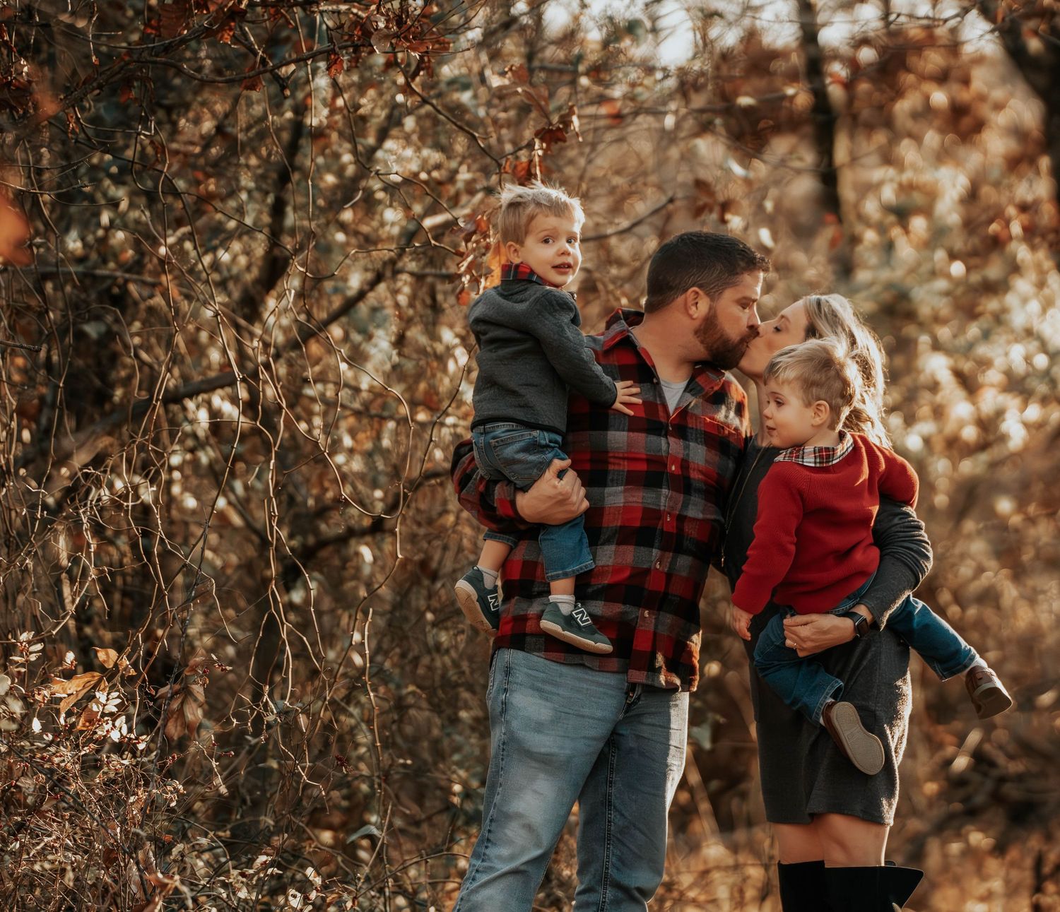 Person in plaid flannel shirt holding two young children in a fall forest setting.