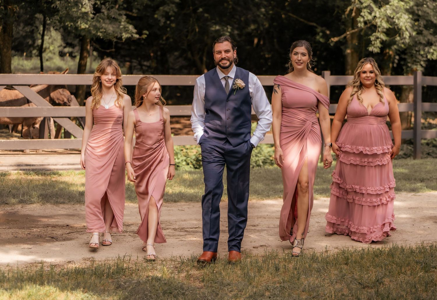 Wedding party members in pink dresses and navy suits walk across a sunny outdoor setting.