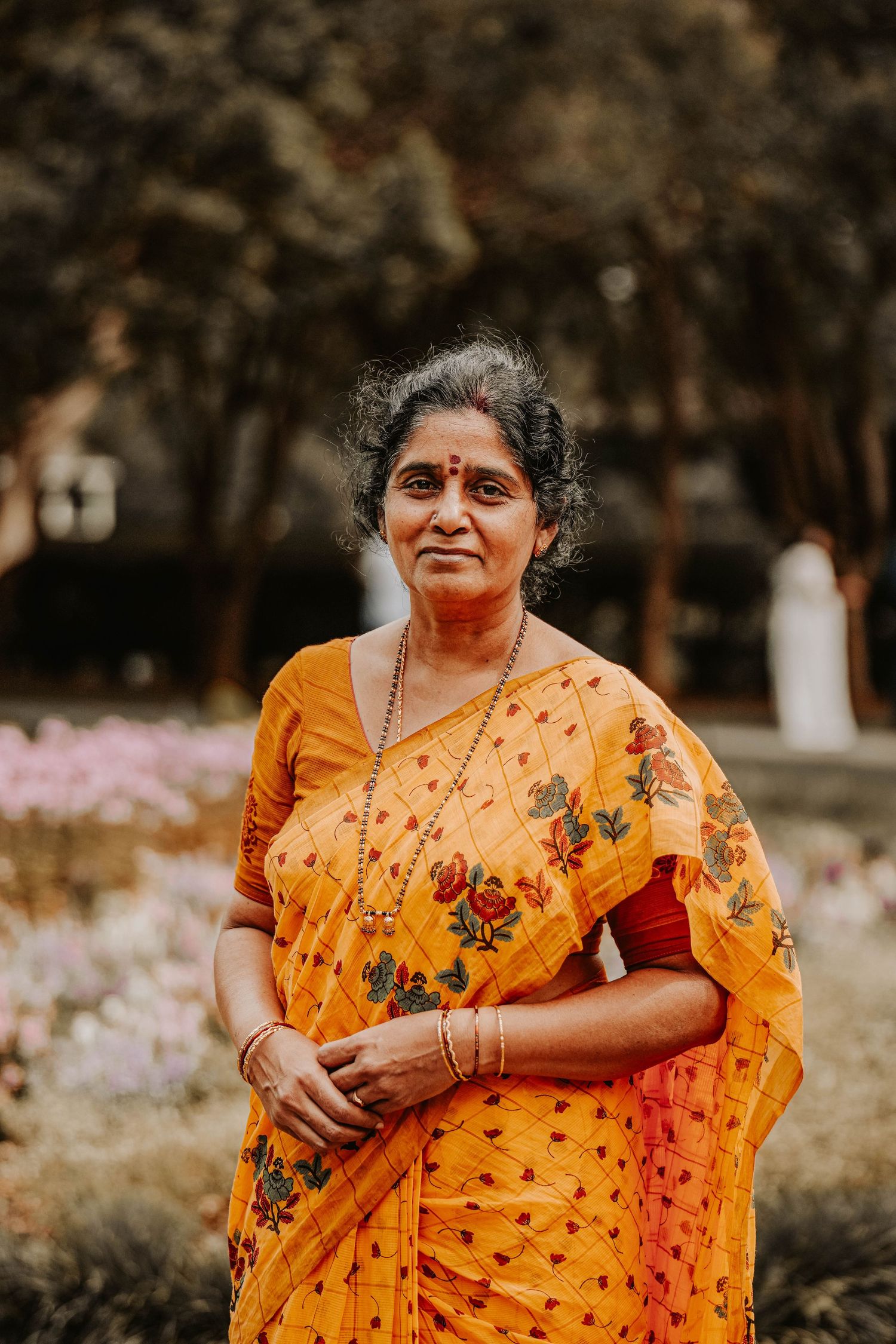 Woman wearing vibrant orange sari poses in a garden setting with blooming flowers.