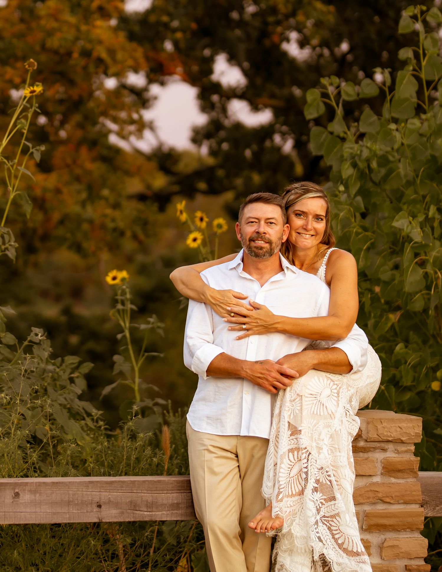 A couple embraces while standing in a rustic outdoor setting with sunflowers and wooden fences at sunset.