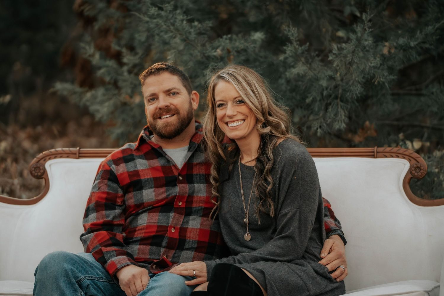 Couple sitting together on white outdoor couch with fall forest backdrop.