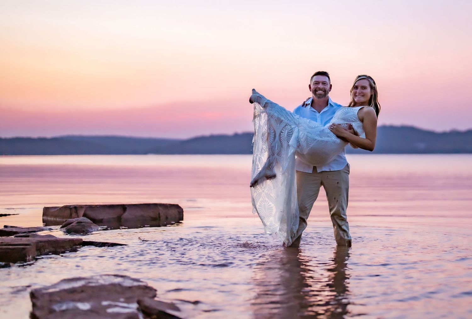 Couple embracing during sunset by the ocean against a pink and purple sky.