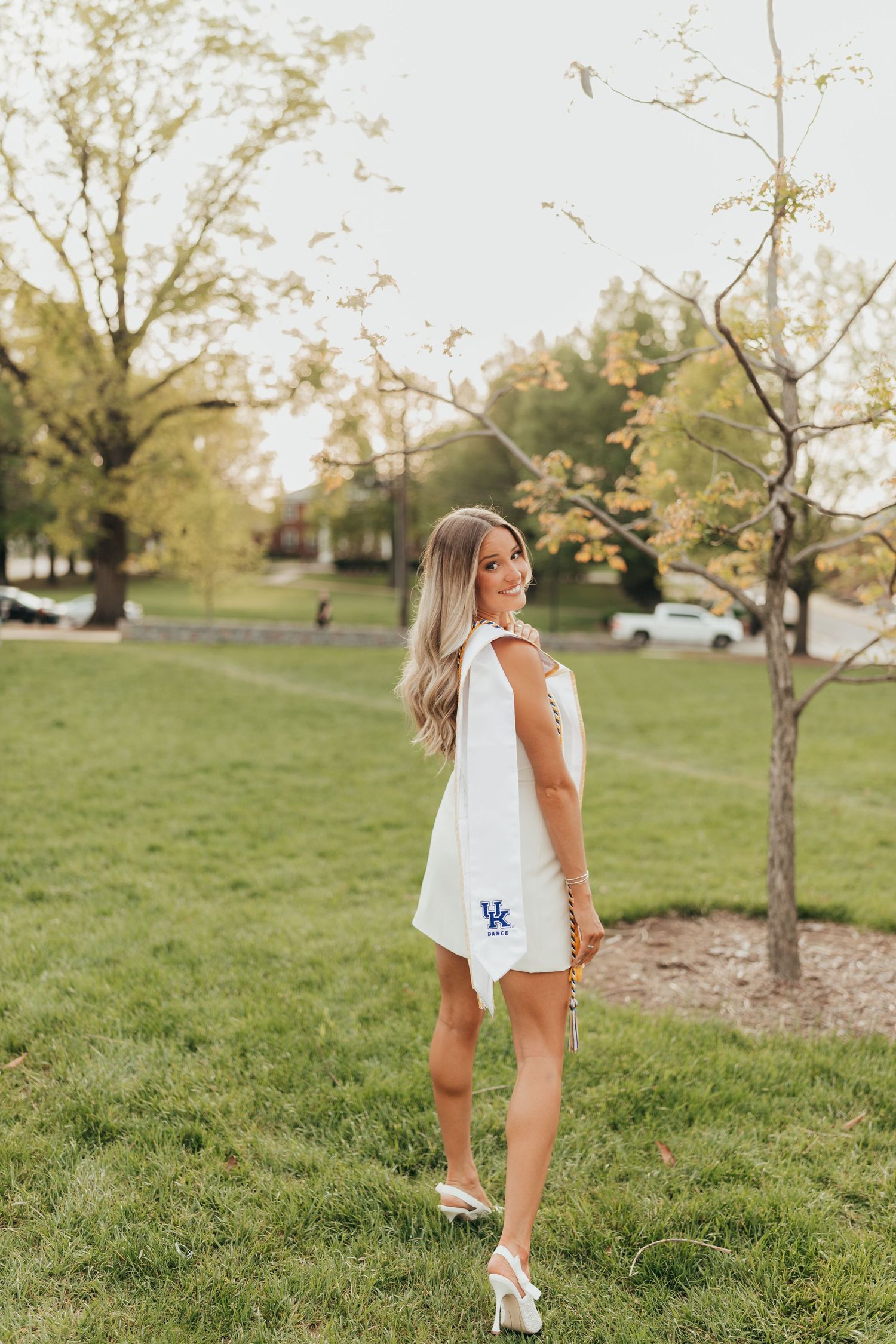 Graduate in white dress poses on grassy campus lawn during golden hour.
