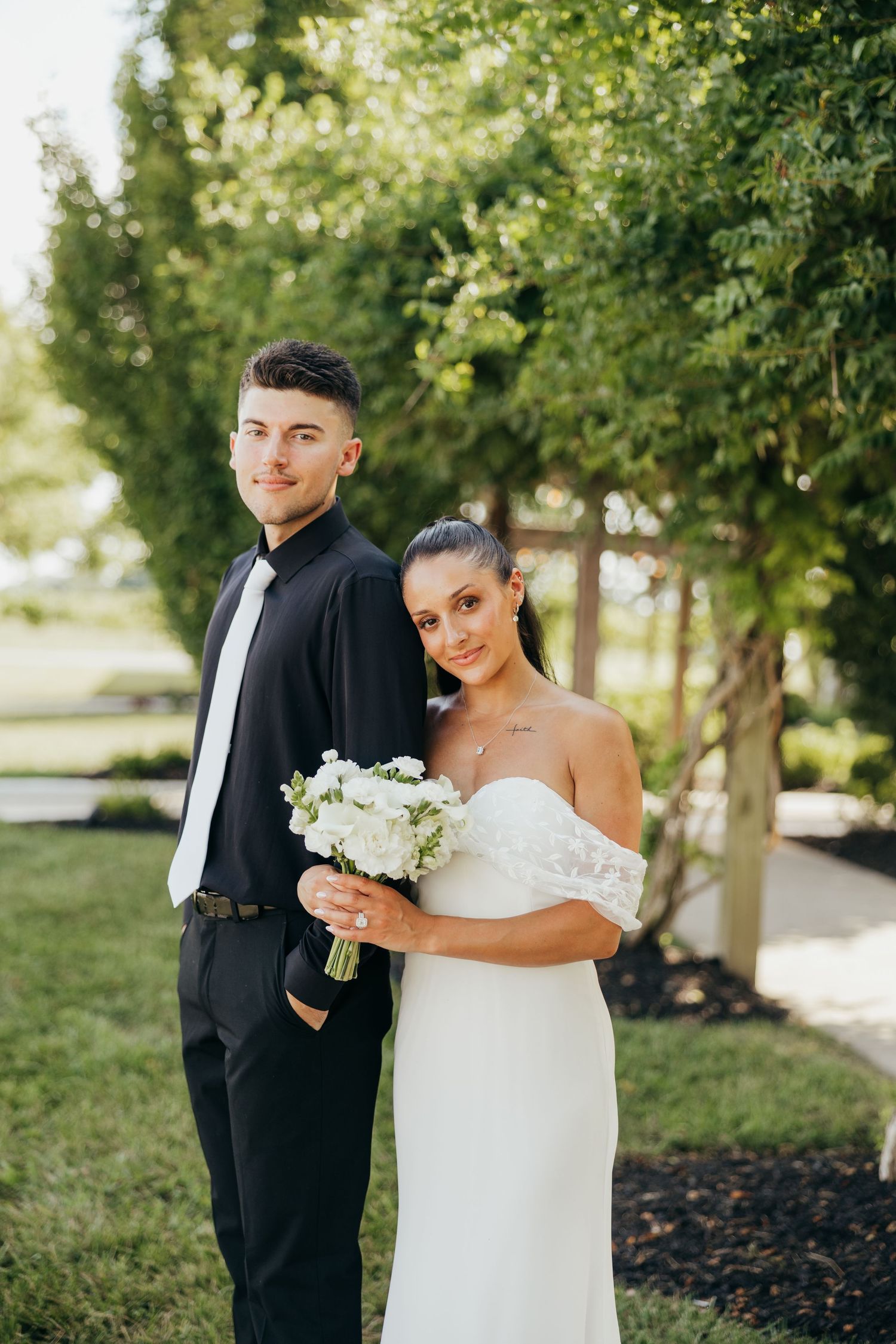 Bride in off-shoulder white gown holding white bouquet standing with groom in black suit outdoors.