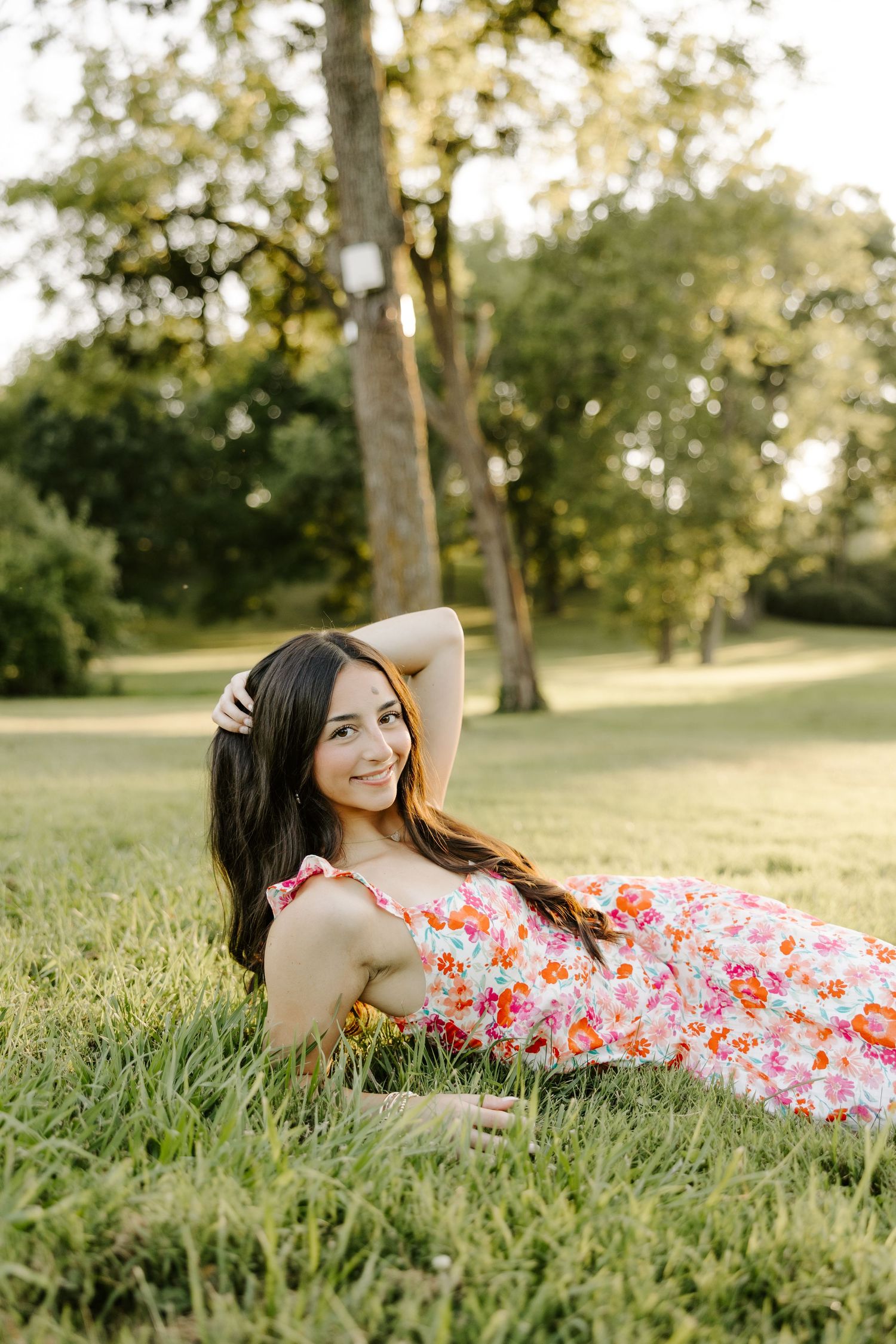 Young woman in floral dress lying on green grass smiling in sunlit park with tall trees.