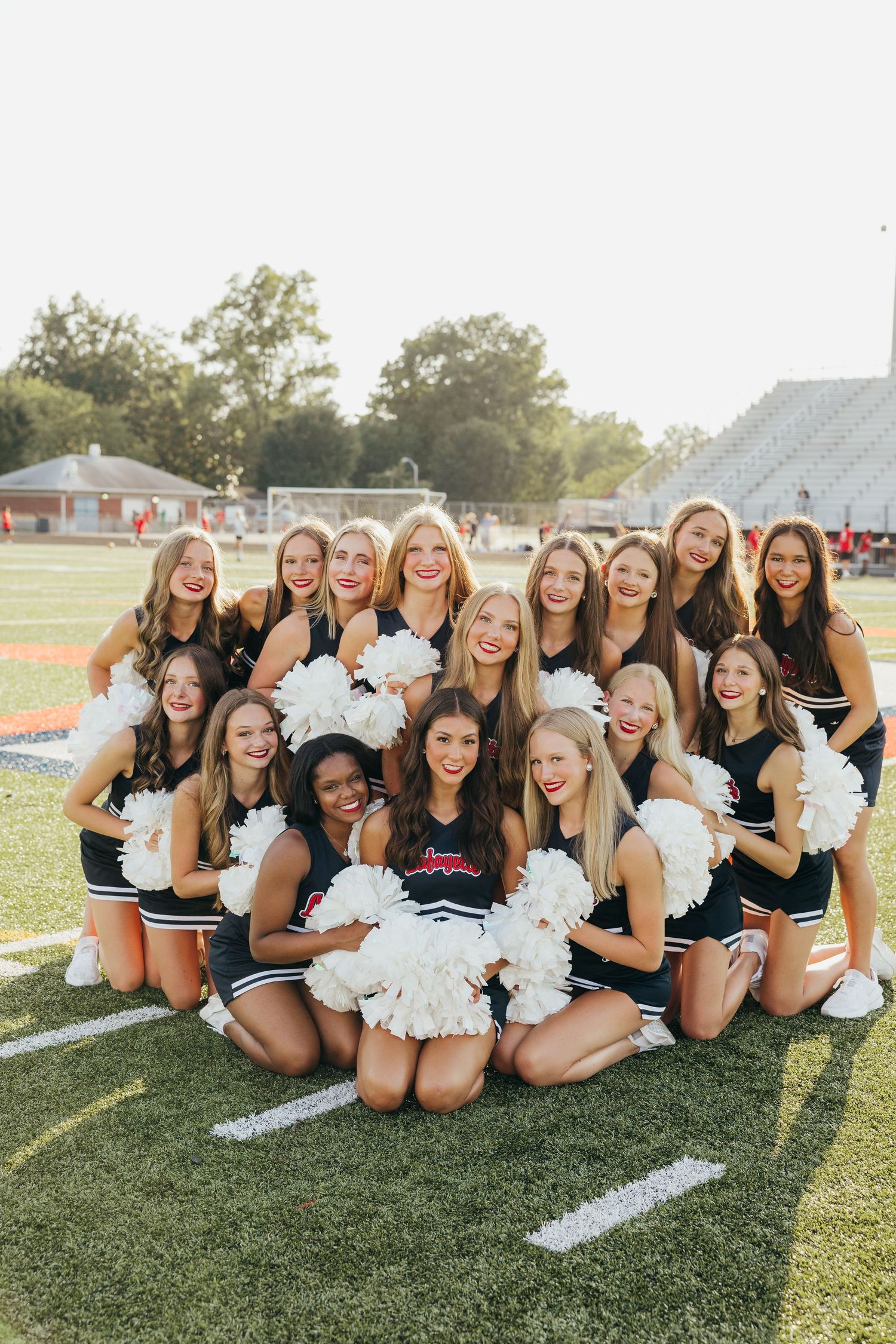 High school cheerleaders in navy uniforms with white pom-poms posing on football field at sunset.