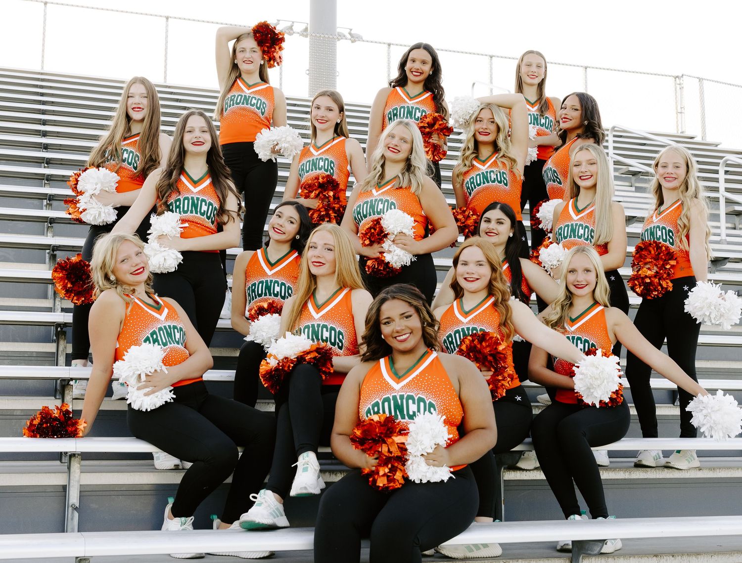 Cheerleading team in orange Broncos uniforms posing with pom-poms on stadium bleachers outdoors.