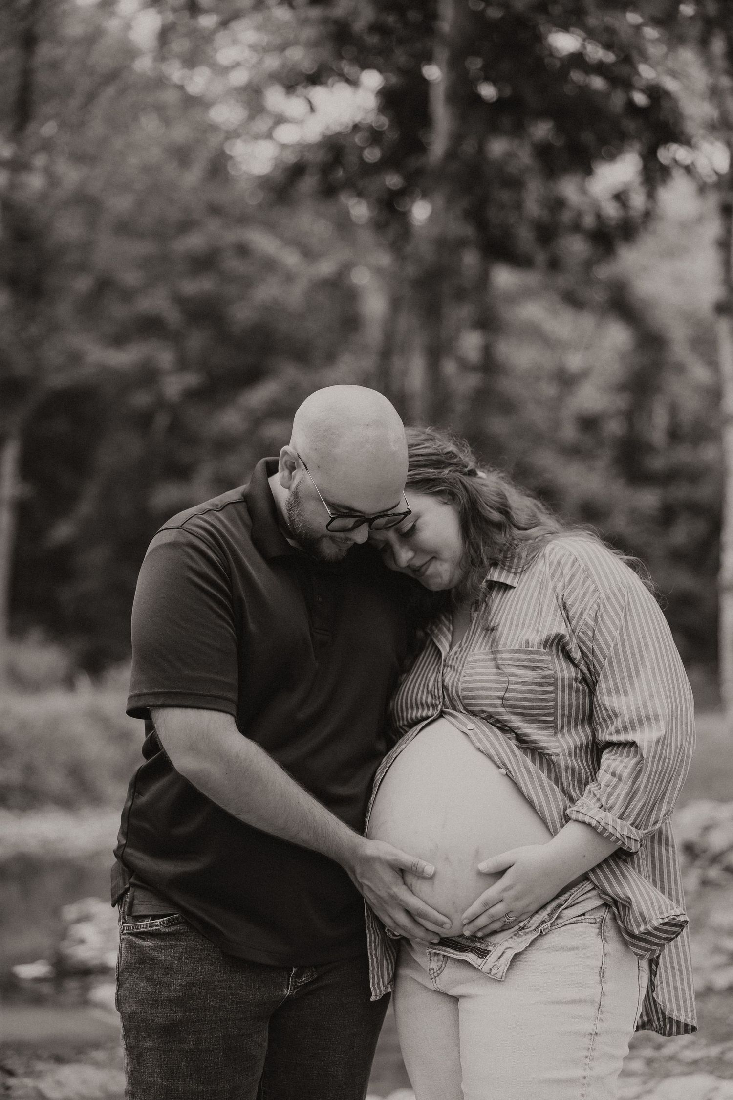 Black and white maternity photo of couple embracing and cradling pregnant belly tenderly in the woods.
