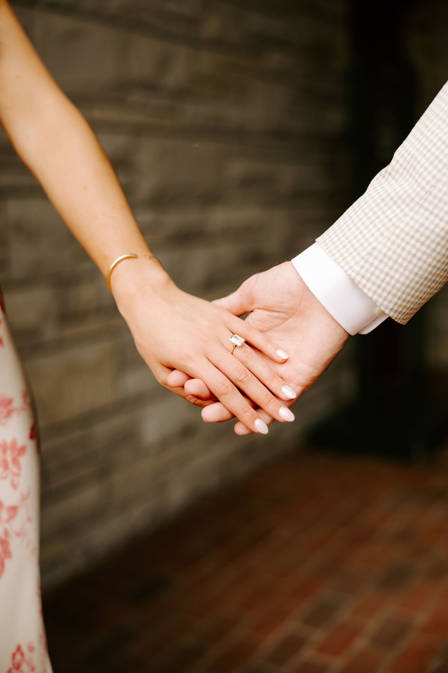 Close-up of couple holding hands showing gold wedding ring and bracelet against stone wall.