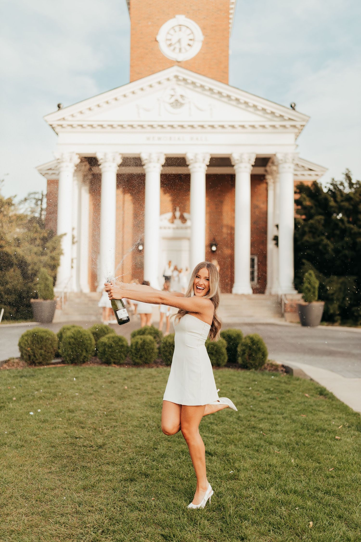 A white summer dress celebration pose in front of a grand neoclassical building with columns and a clock tower.