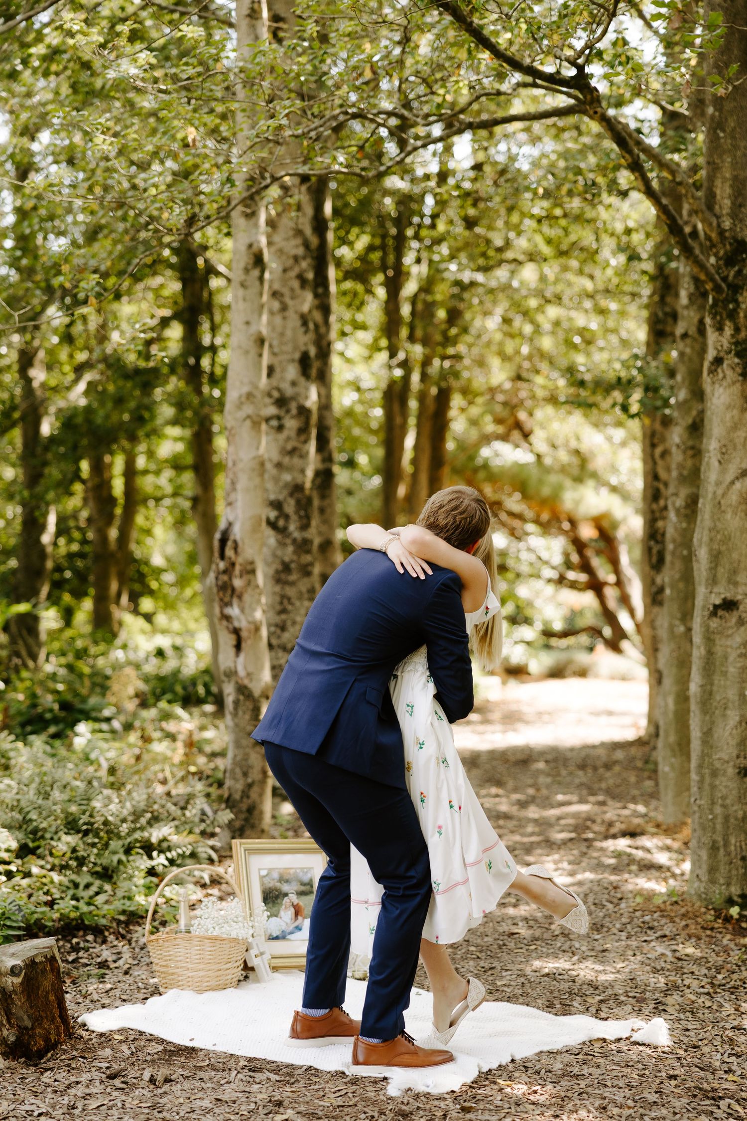 An emotional embrace after a proposal on a wooded path with picnic setup.