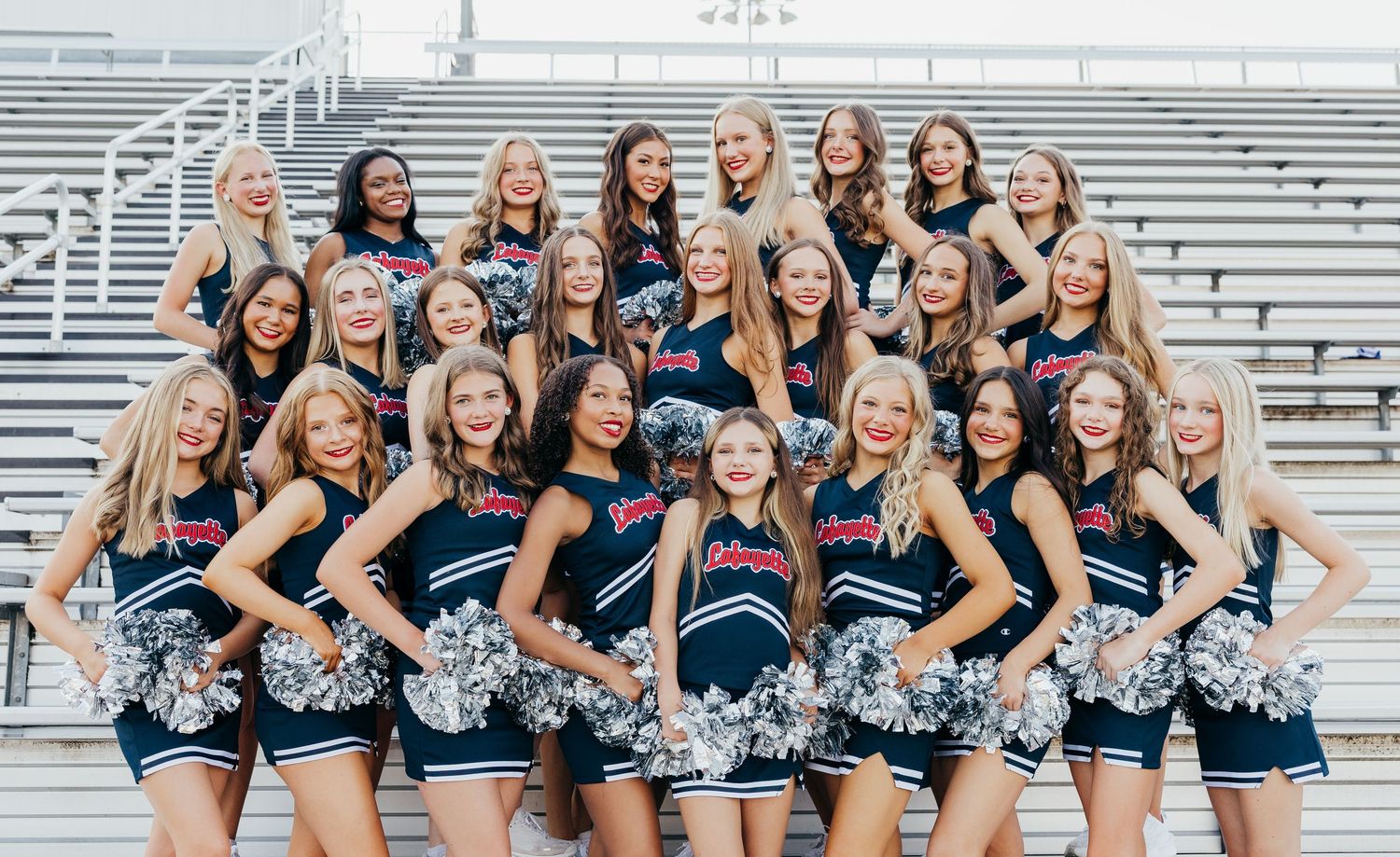 Large cheerleading team in navy uniforms with silver pom-poms posing on stadium bleachers outdoors.