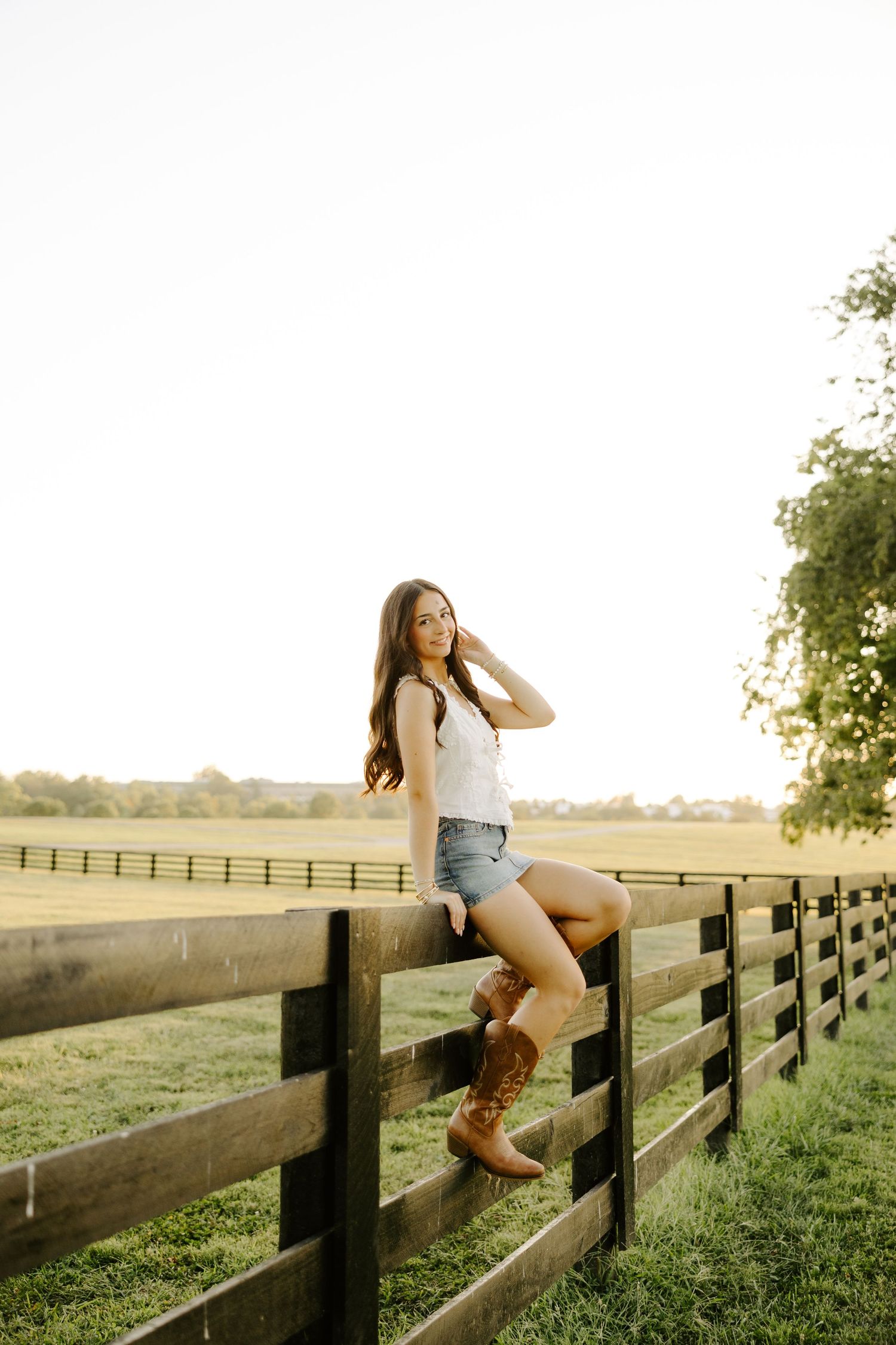 Young woman in denim shorts and cowboy boots sitting on a rustic wooden farm fence at golden hour.