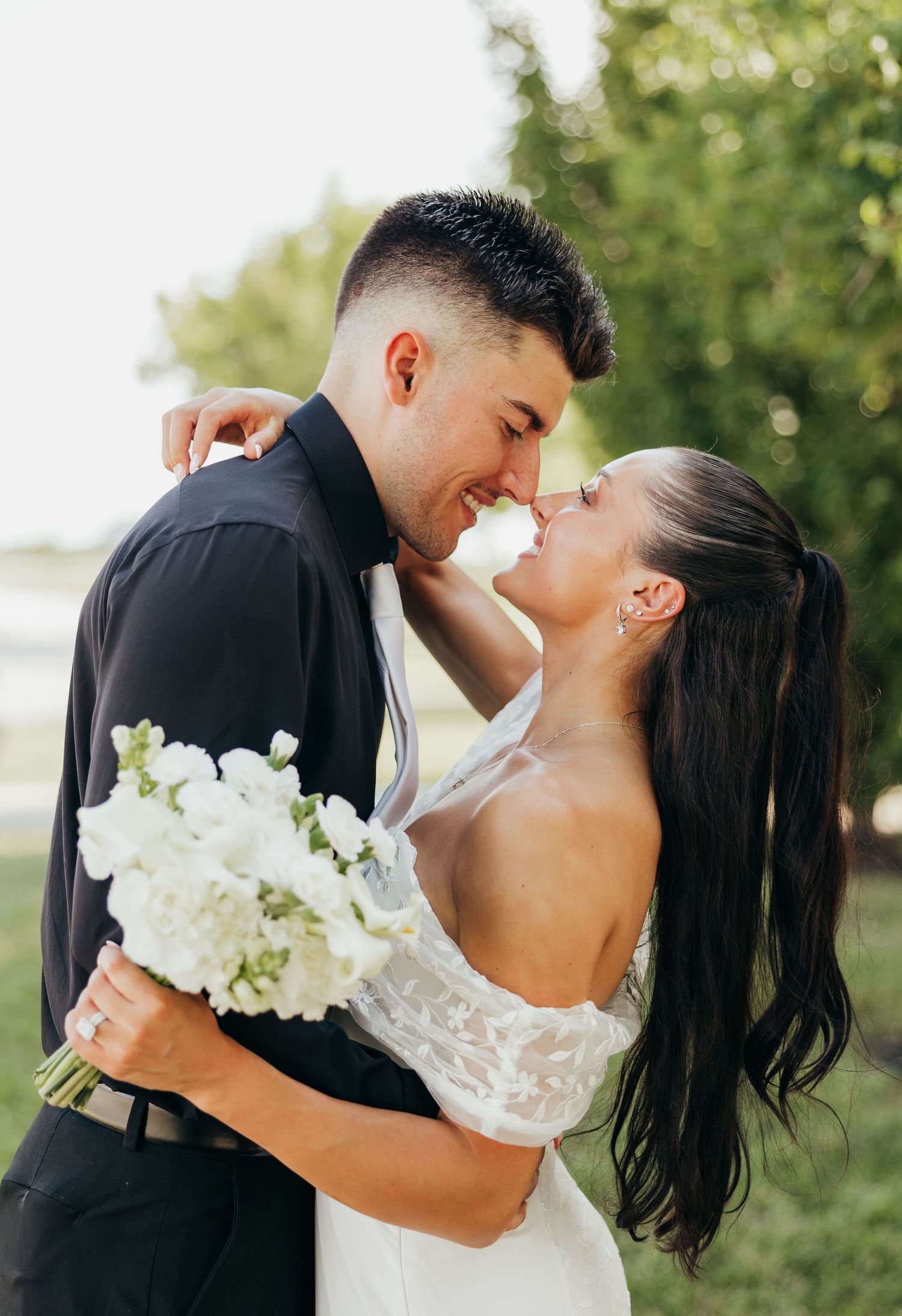 Bride and groom sharing romantic nose-to-nose moment while bride holds white floral bouquet outdoors.