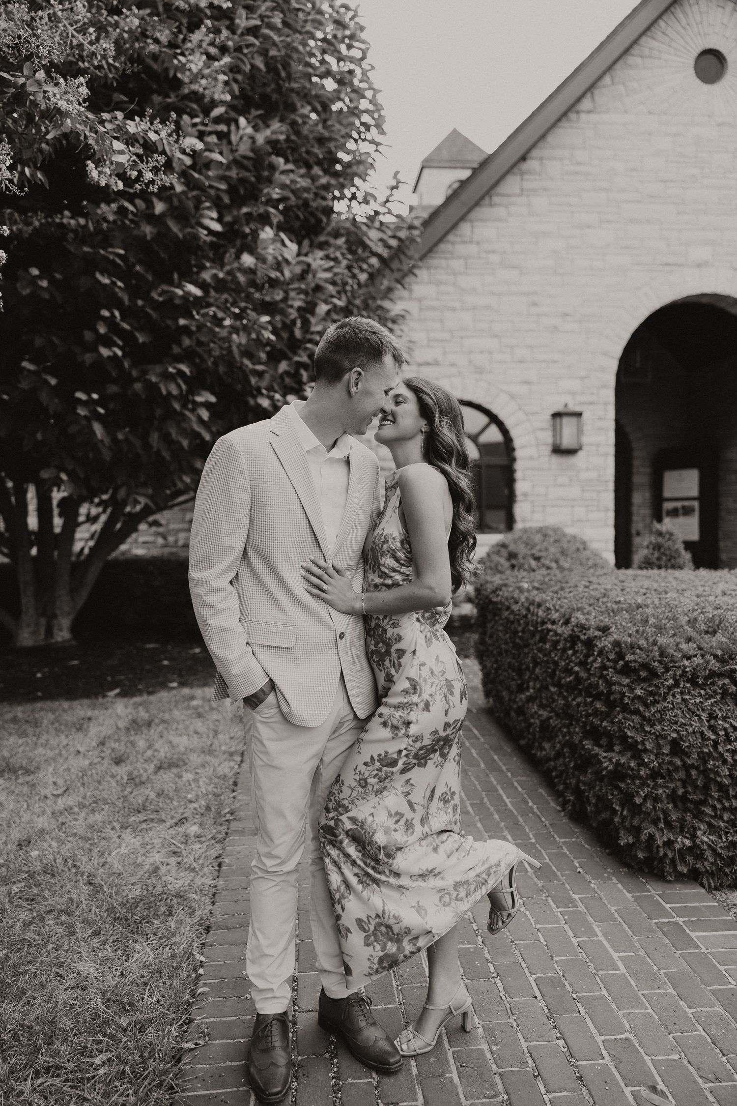 Black and white photo of a couple laughing and embracing on a brick pathway outside a stone building.