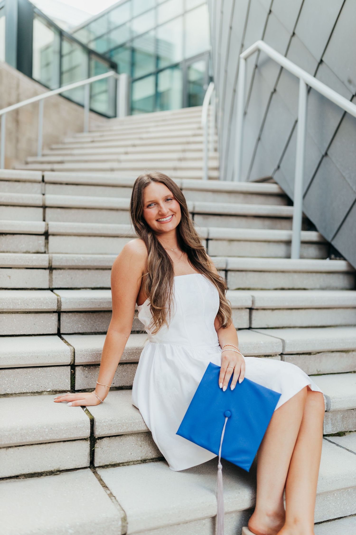 Graduate in white dress sits on concrete steps with blue diploma holder.
