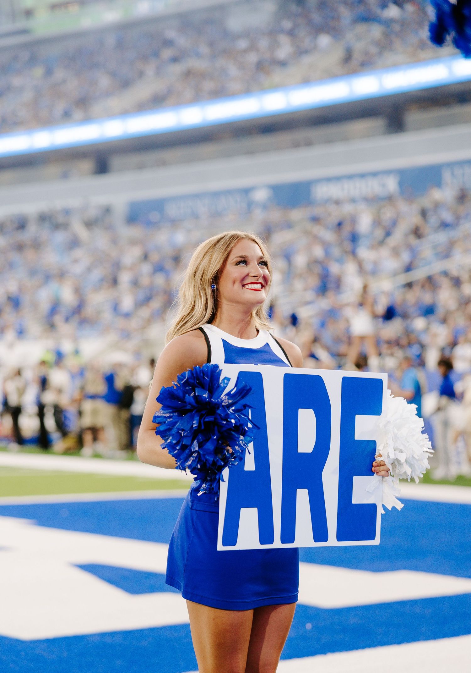 A smiling UK cheerleader holds a sign reading ARE with blue pom-poms during a Kentucky Wildcats football game.