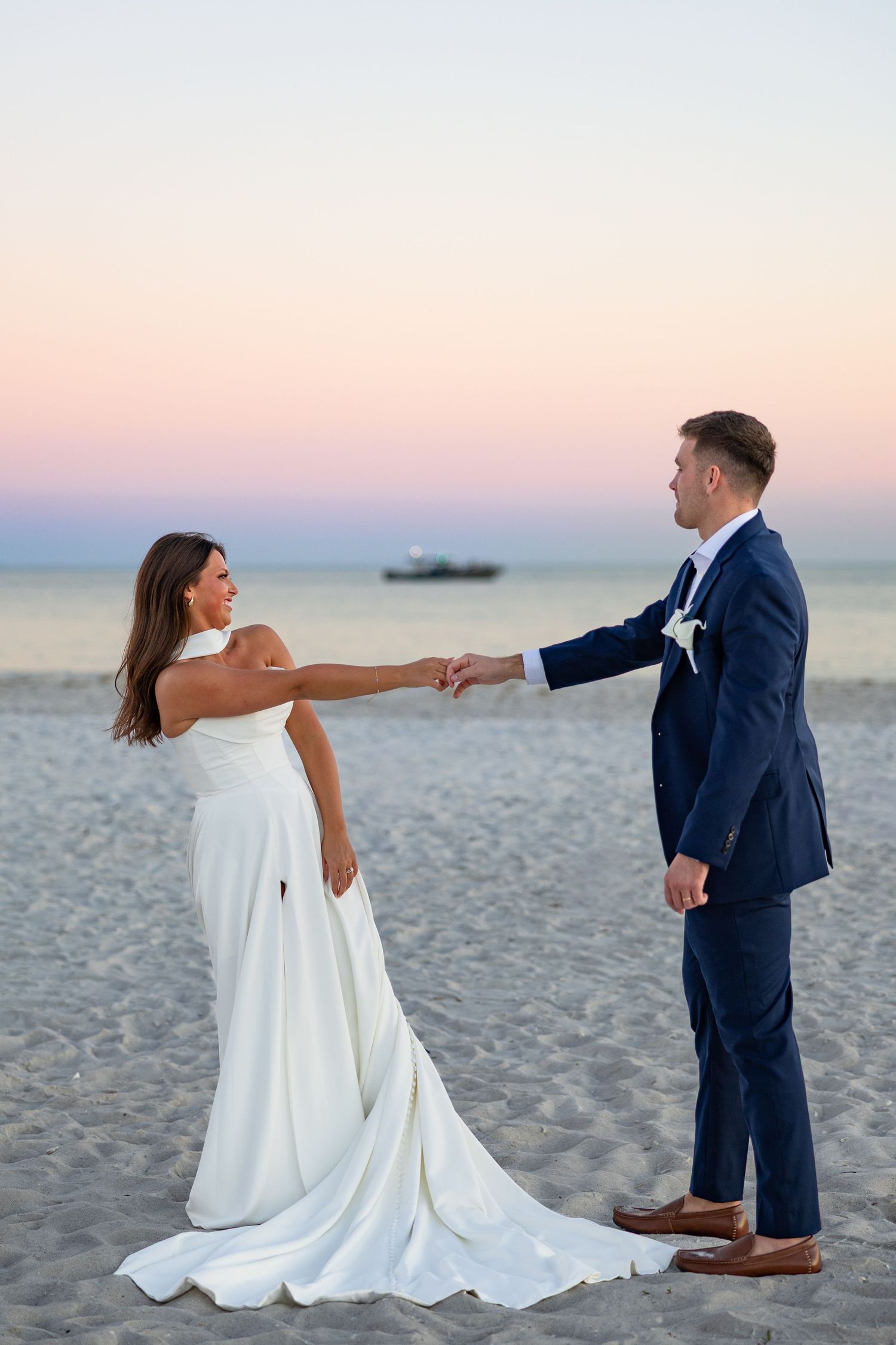 A bride in a white gown and groom in navy suit hold hands on a sandy beach at sunset.
