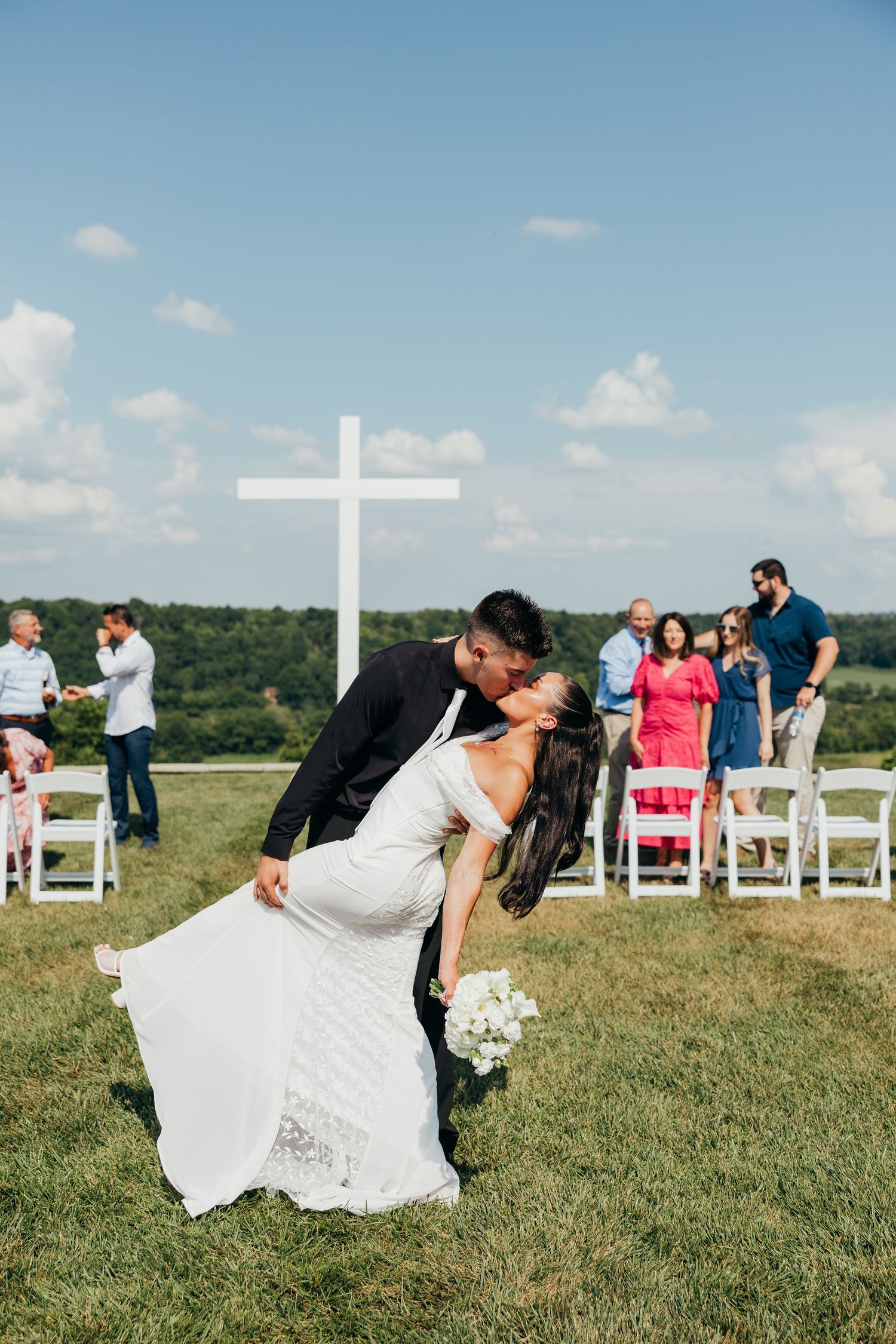 Groom dips bride for a kiss after outdoor wedding ceremony with guests and white cross in background.