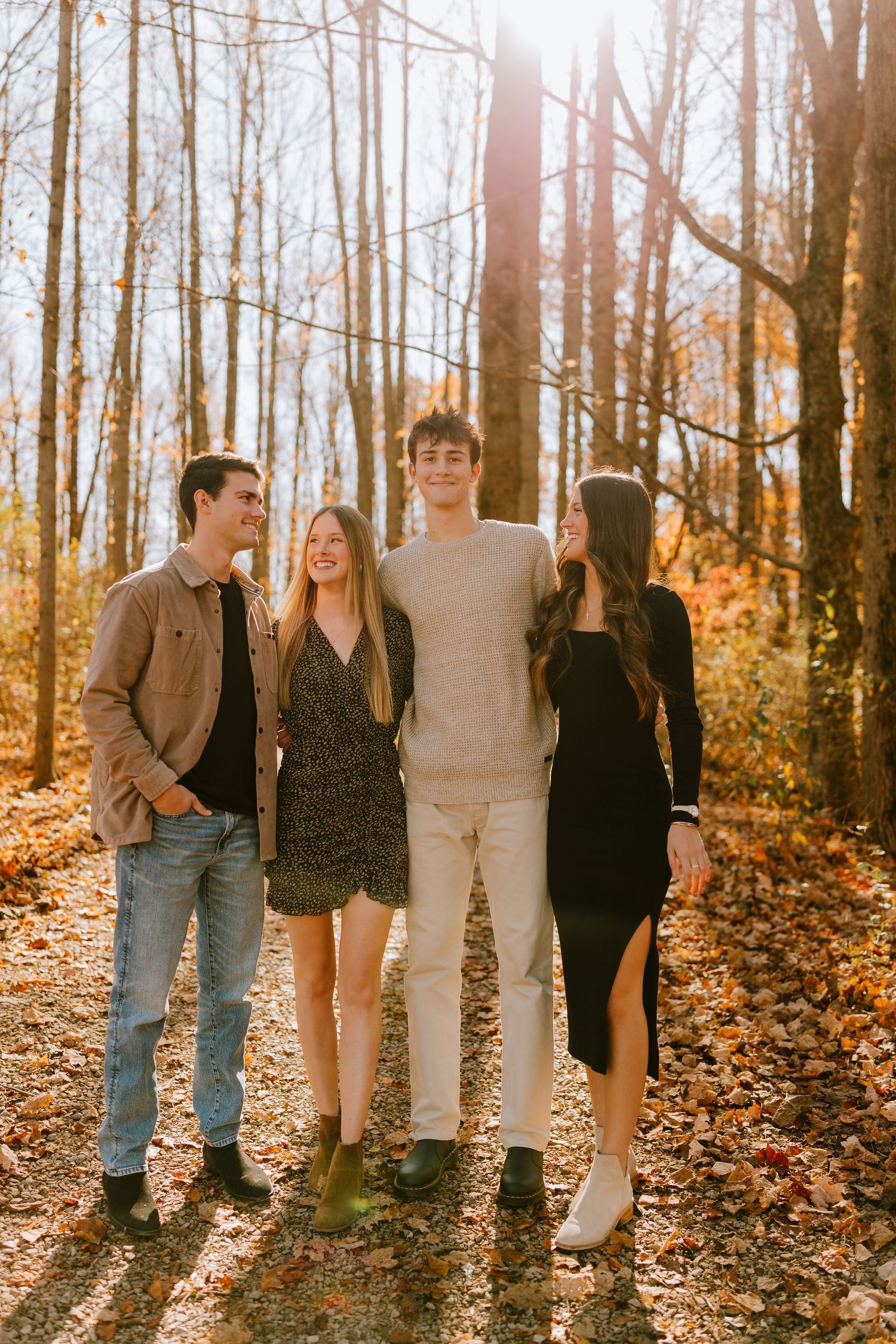 Four young adults smiling and walking together on a leaf-covered forest path during autumn.