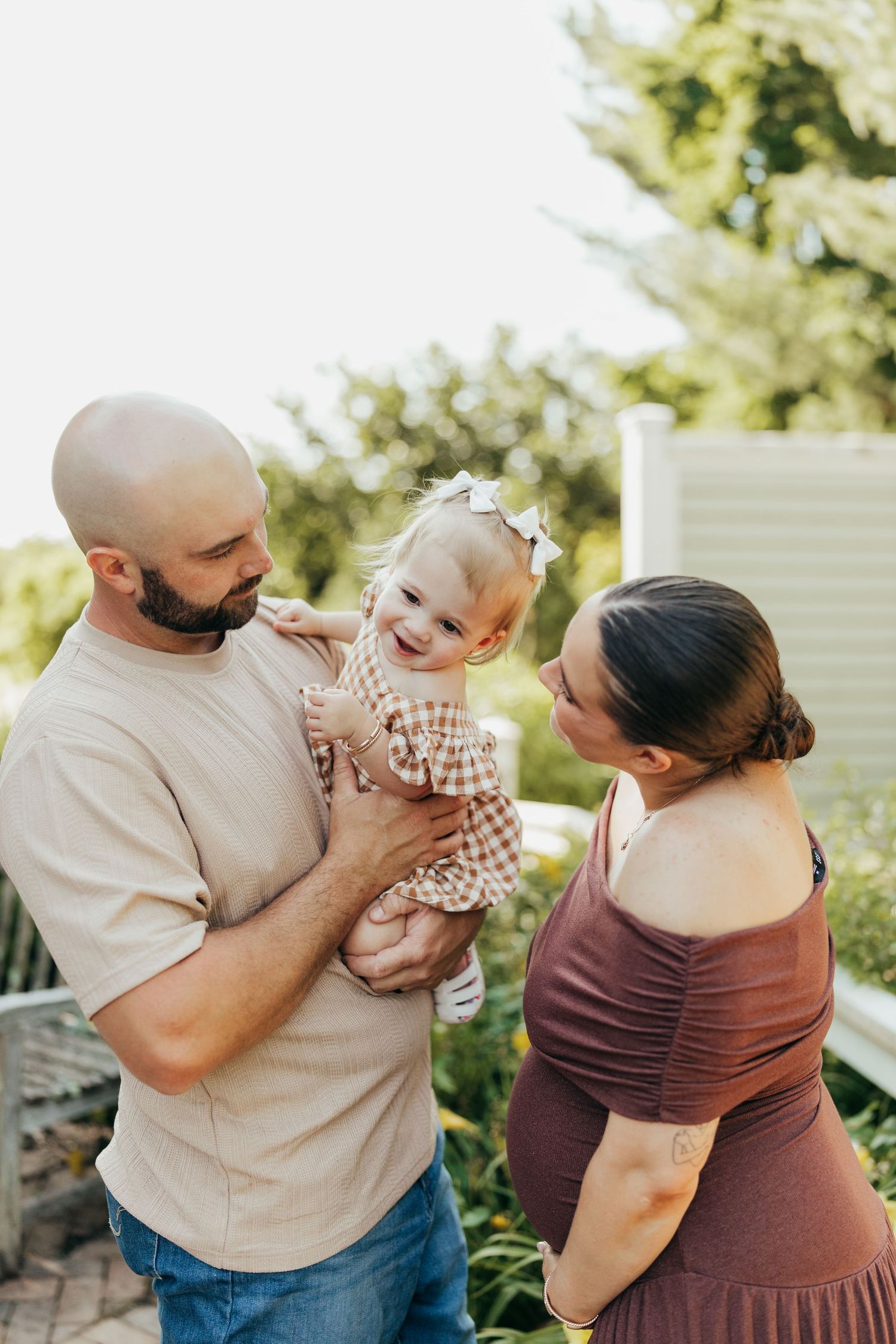 Bald father holding laughing toddler daughter while pregnant mother smiles in sunny backyard.