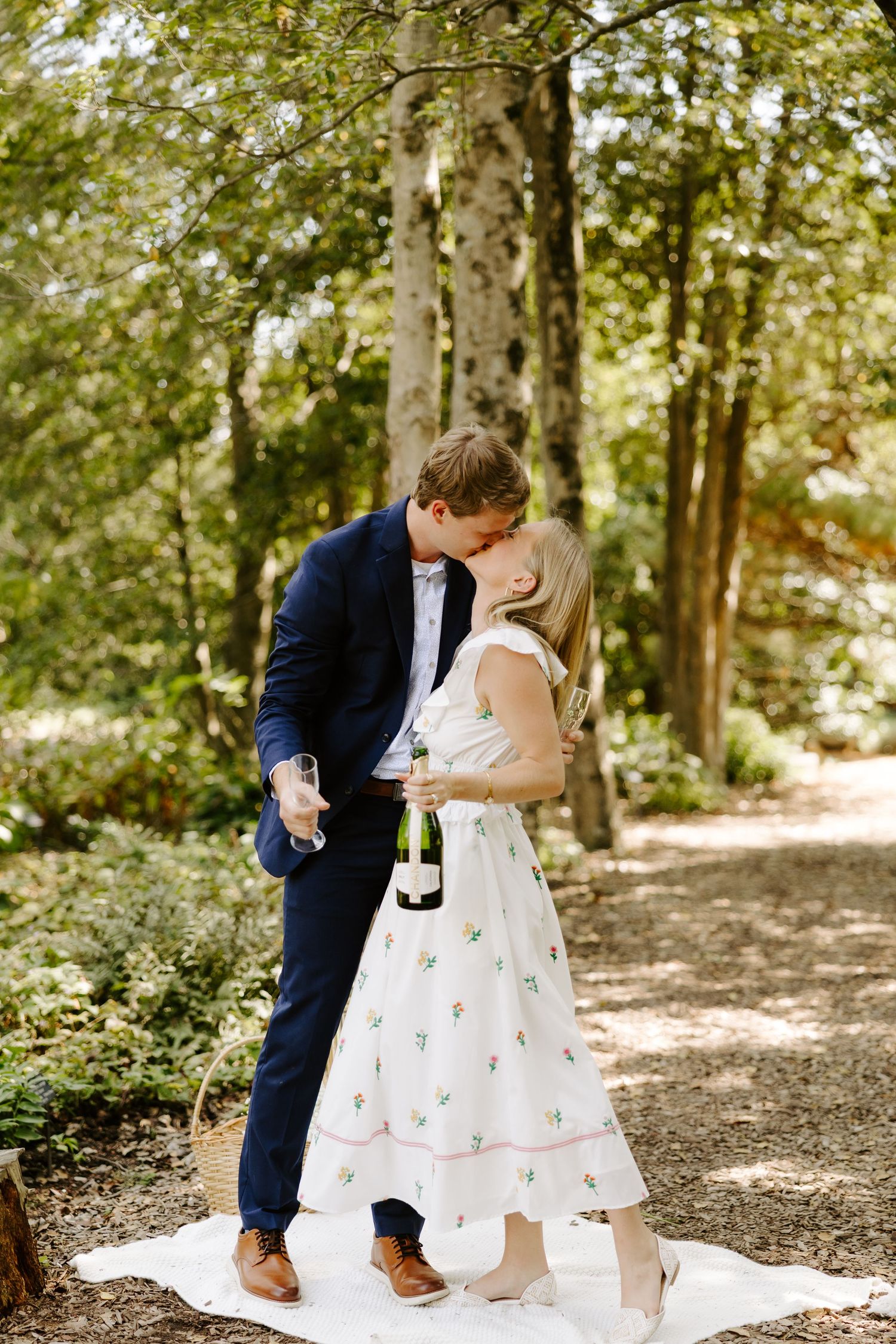 Couple sharing champagne on a picnic blanket in a sunlit forest setting.
