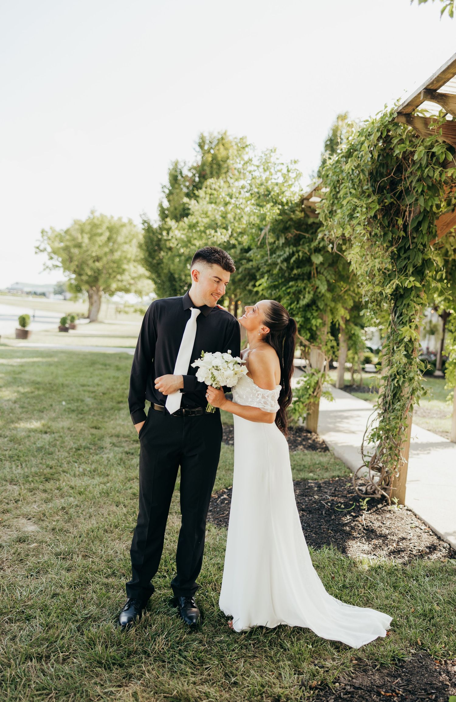Bride in lace wedding gown kissing groom on cheek near ivy-covered pergola at outdoor venue.
