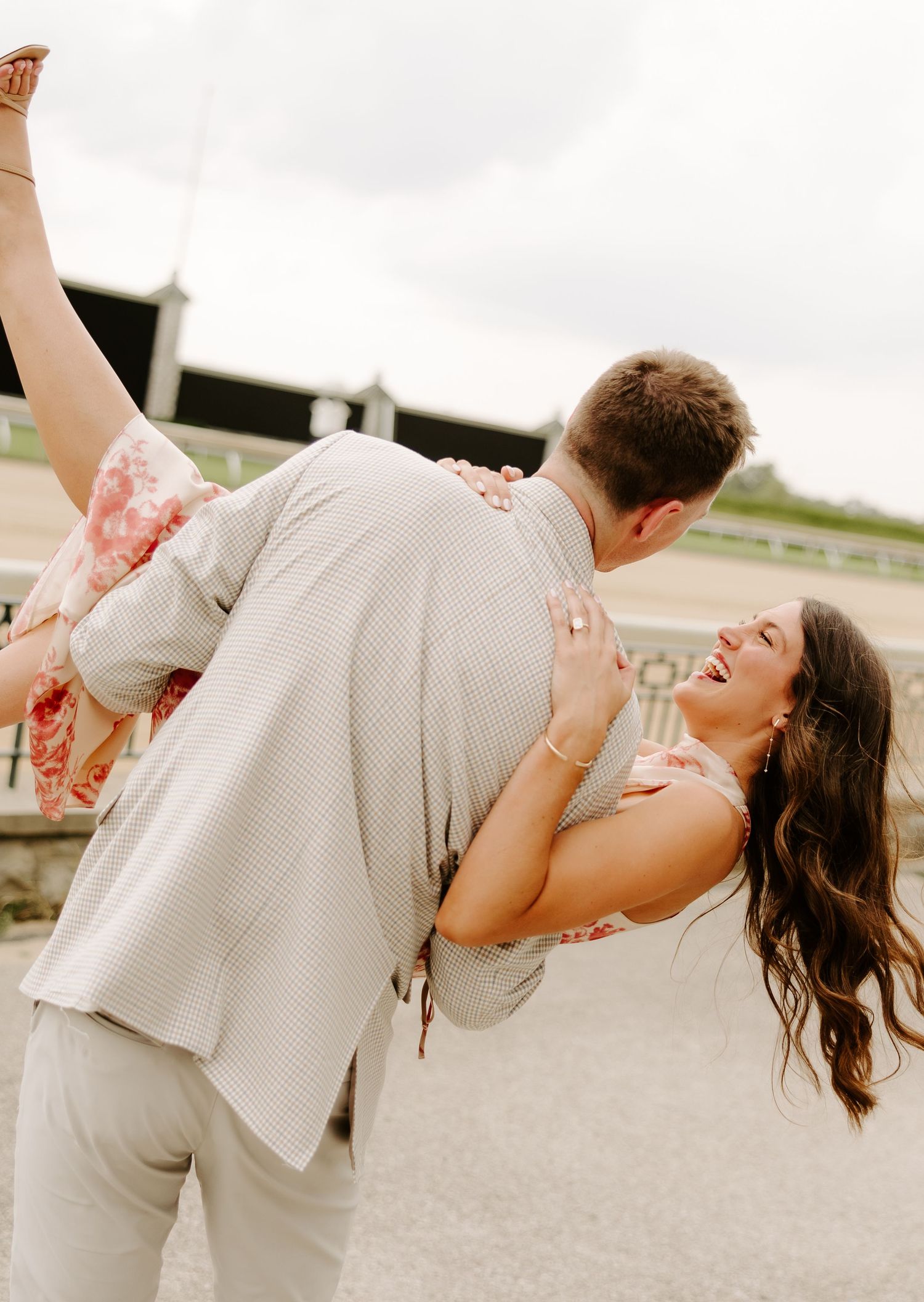 Groom lifts laughing bride in floral dress outdoors at engagement photo session.
