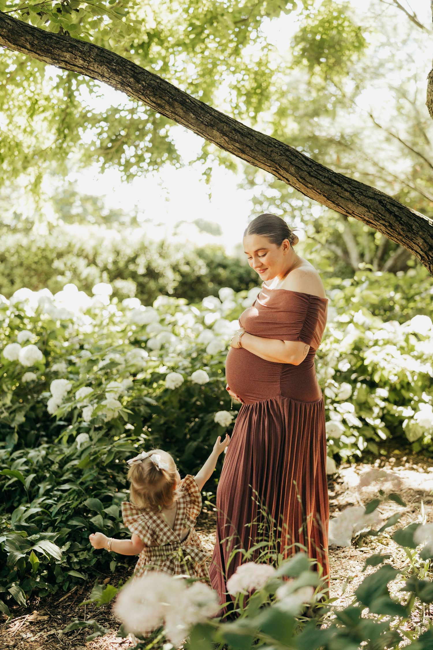 Pregnant woman in burgundy dress holding belly while toddler daughter reaches up in flower garden.