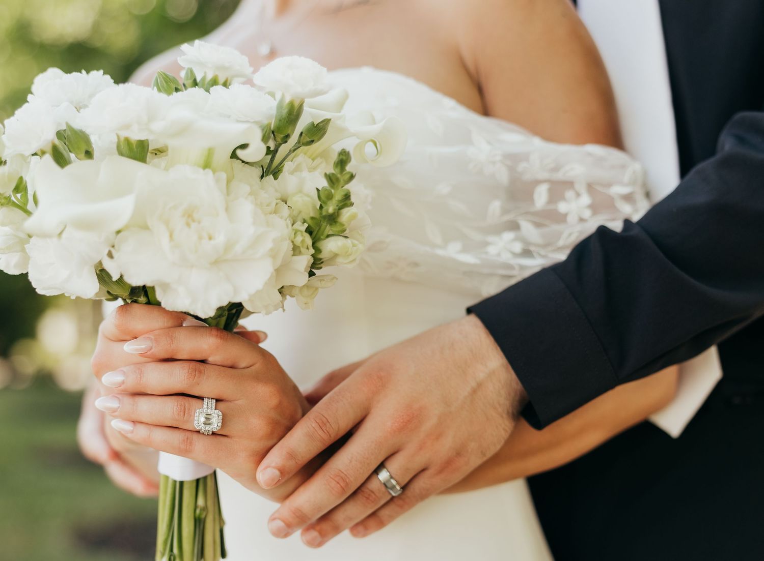 Close-up of bride and groom hands showing diamond wedding rings with white floral bouquet.