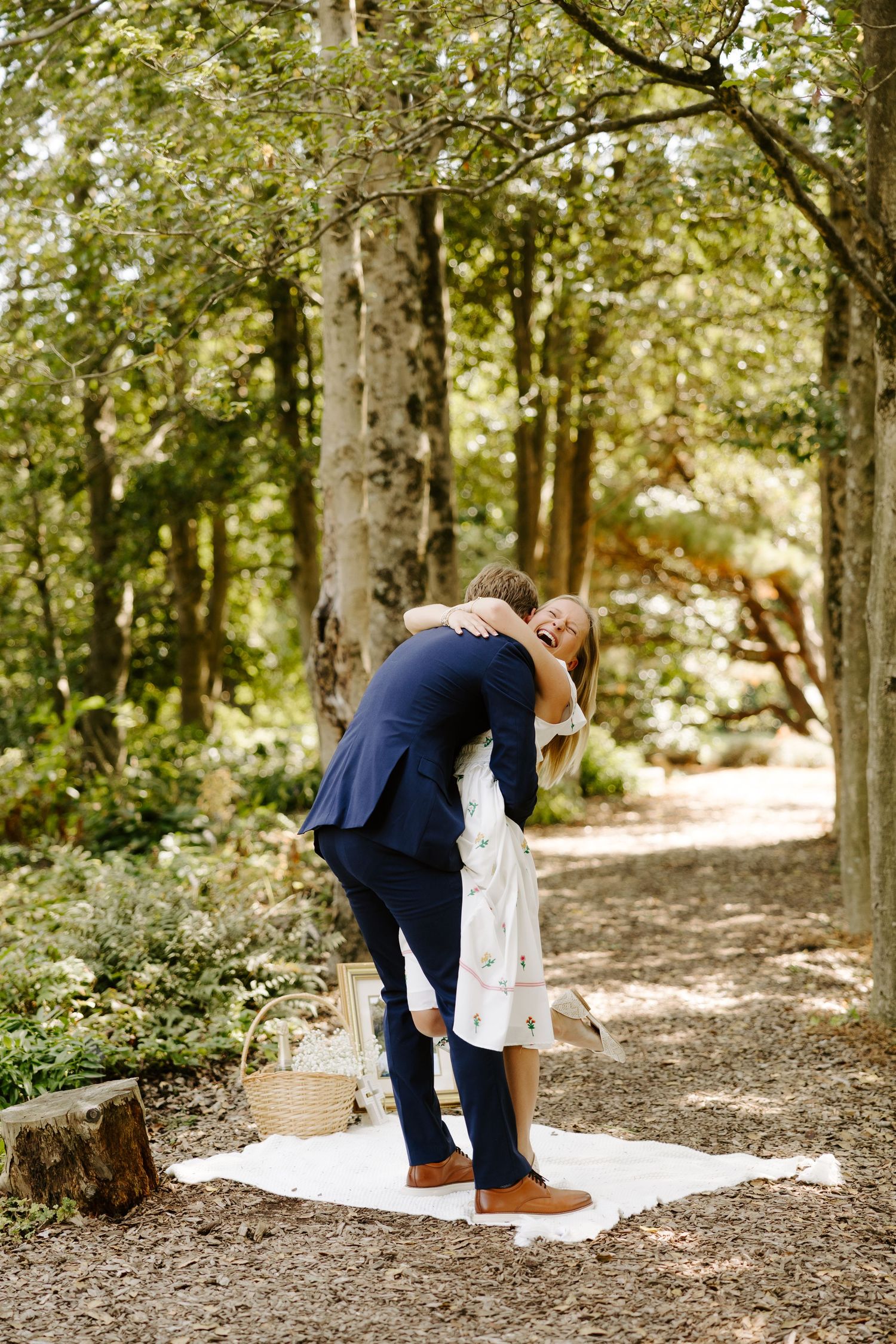 A romantic picnic scene in a wooded area with a white blanket spread on the ground.