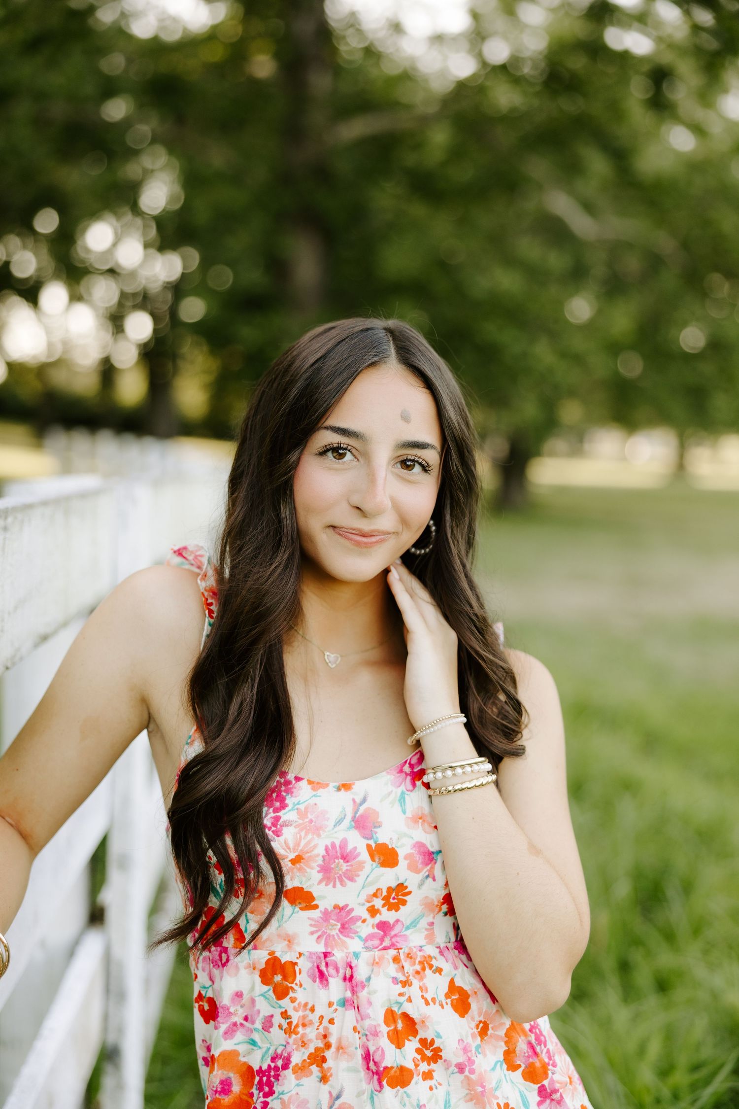 Beautiful brunette teen girl in colorful floral dress posing by white fence in green park.