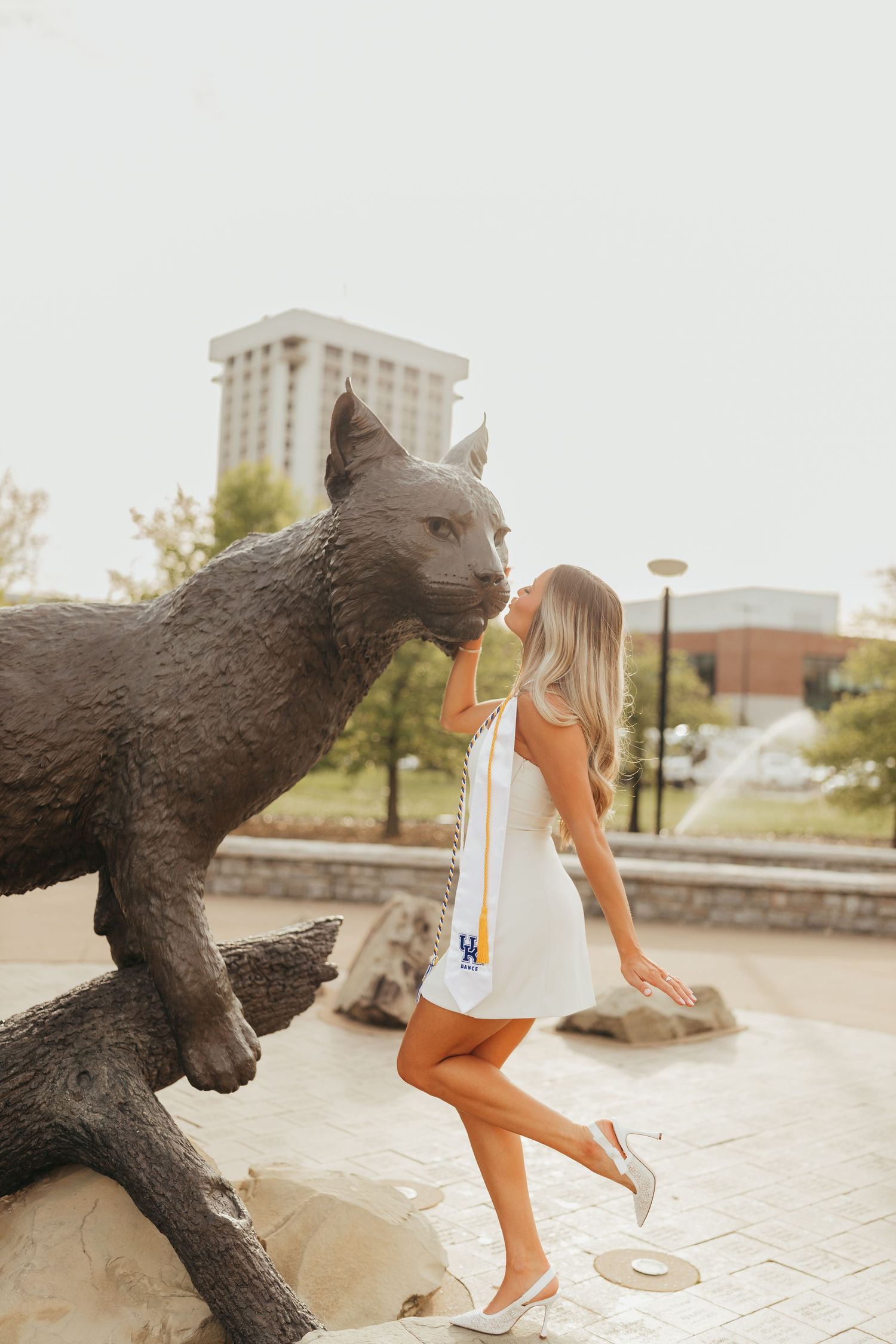 Graduate in white dress poses energetically beside large bronze wildcat statue.