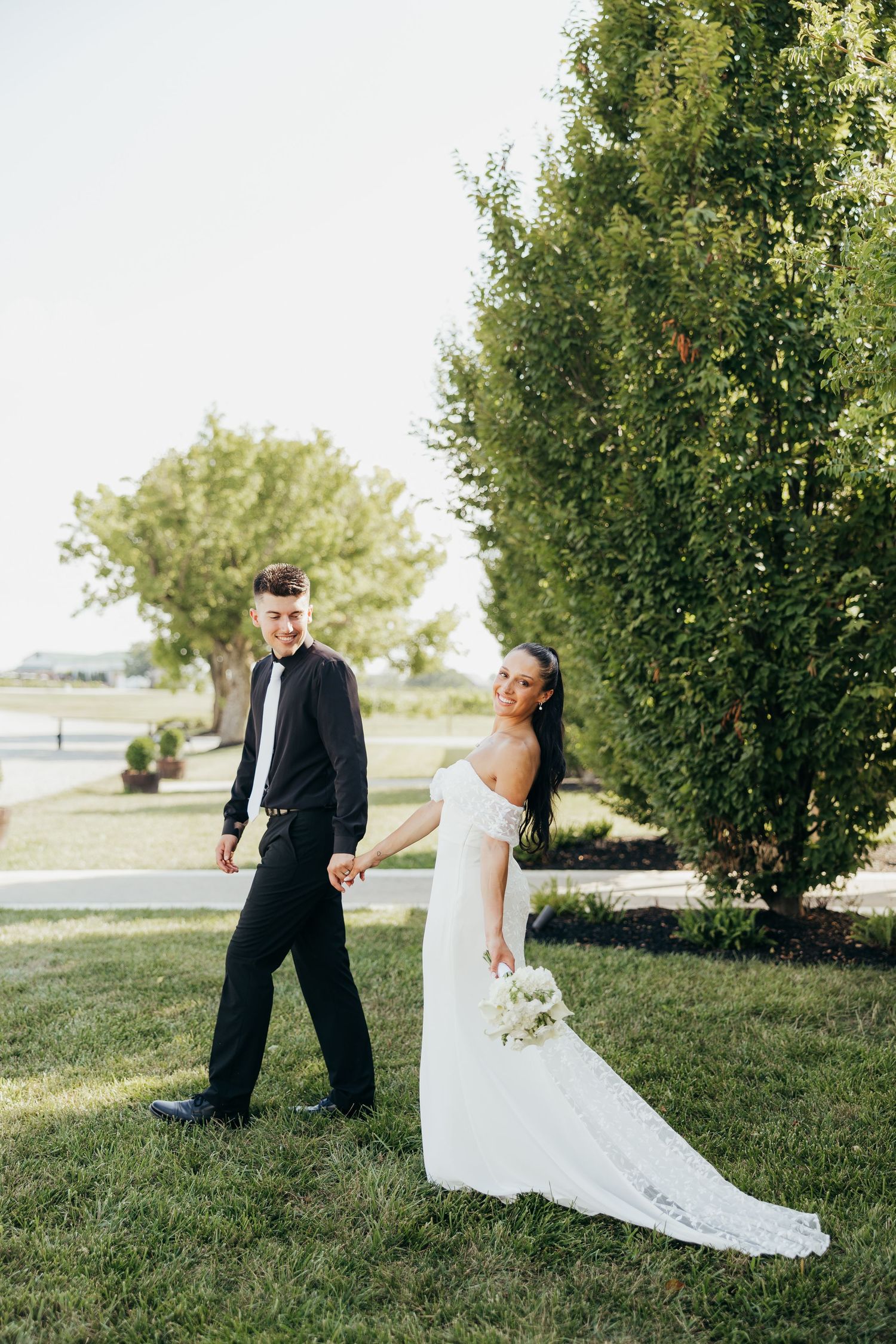 Bride and groom holding hands walking through lush green outdoor venue on wedding day.