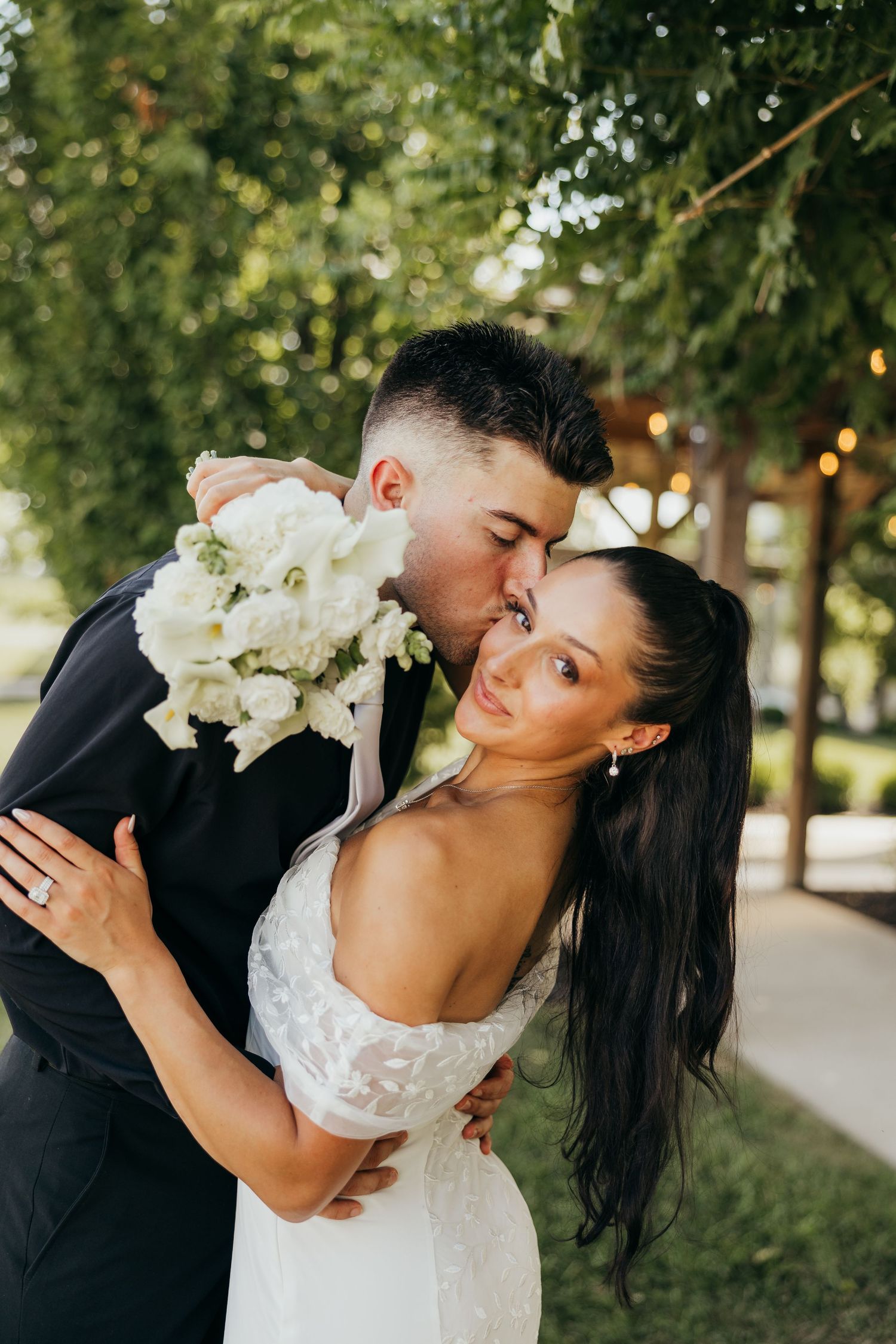 Groom tenderly kisses bride on the temple as she holds a white bouquet under green garden trees.