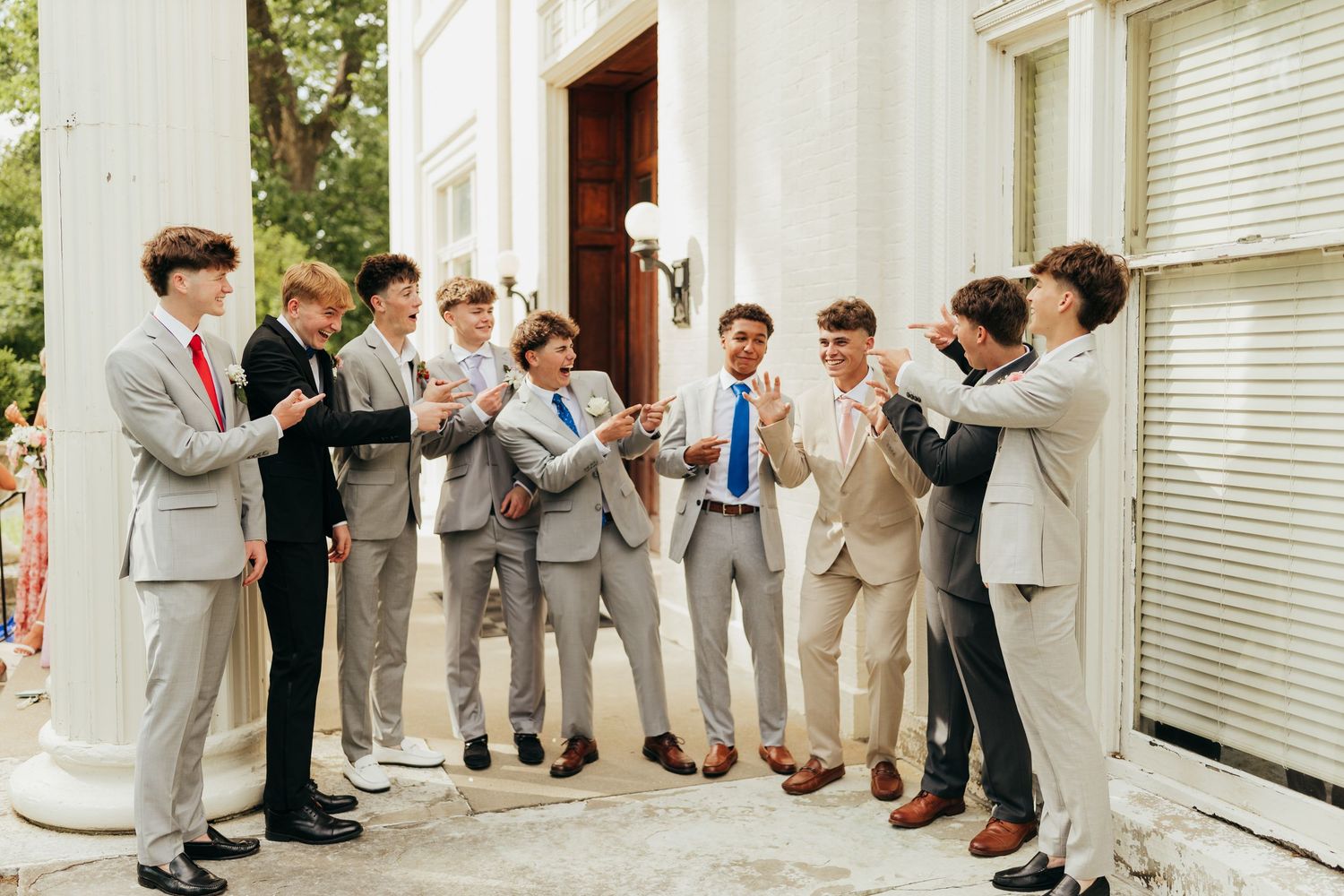 Group of eight young men in grey and beige suits laughing and clapping outside a white building.